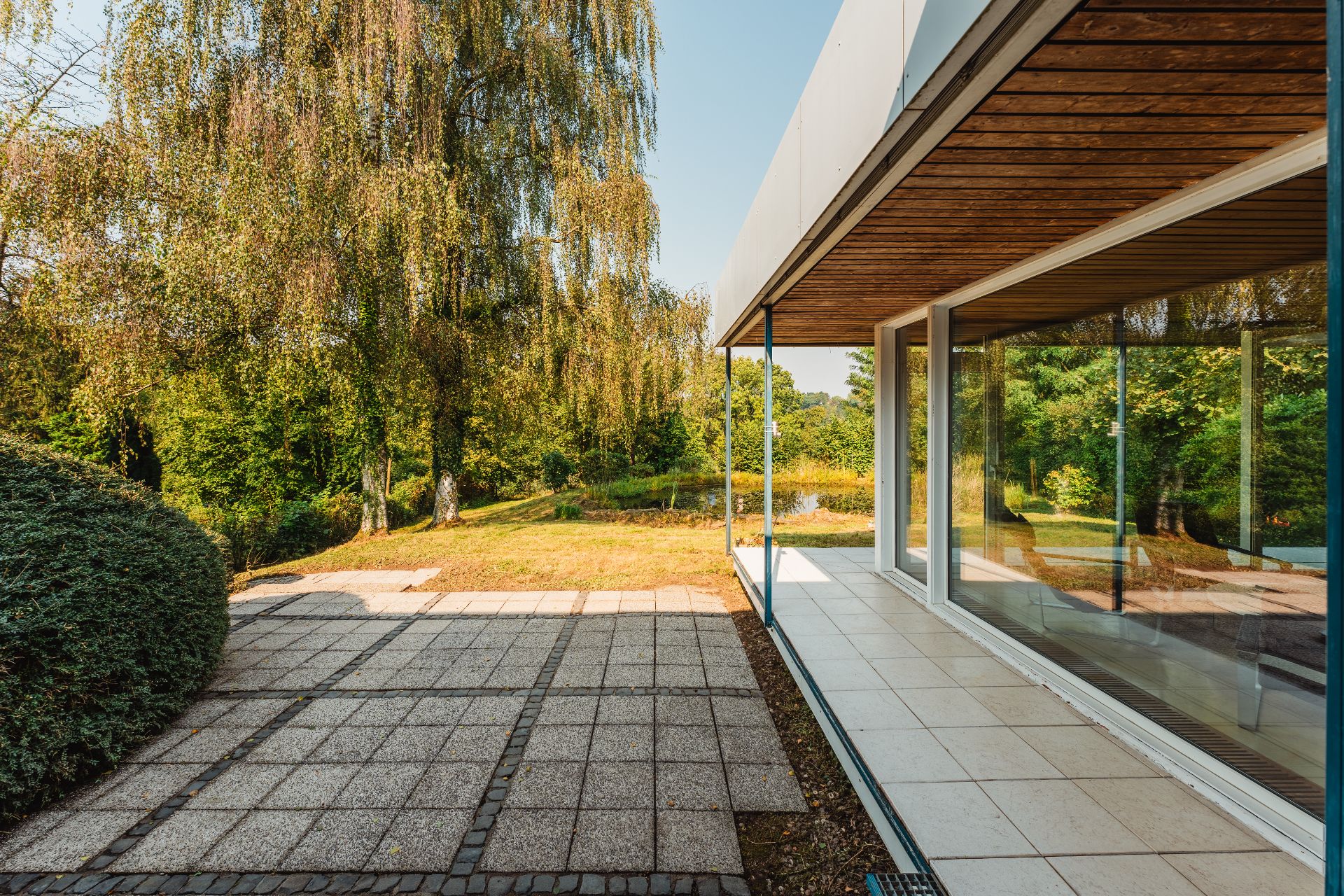 Moderne Terrasse mit großen Glasfenstern und Blick auf einen grünen Garten mit Bäumen.