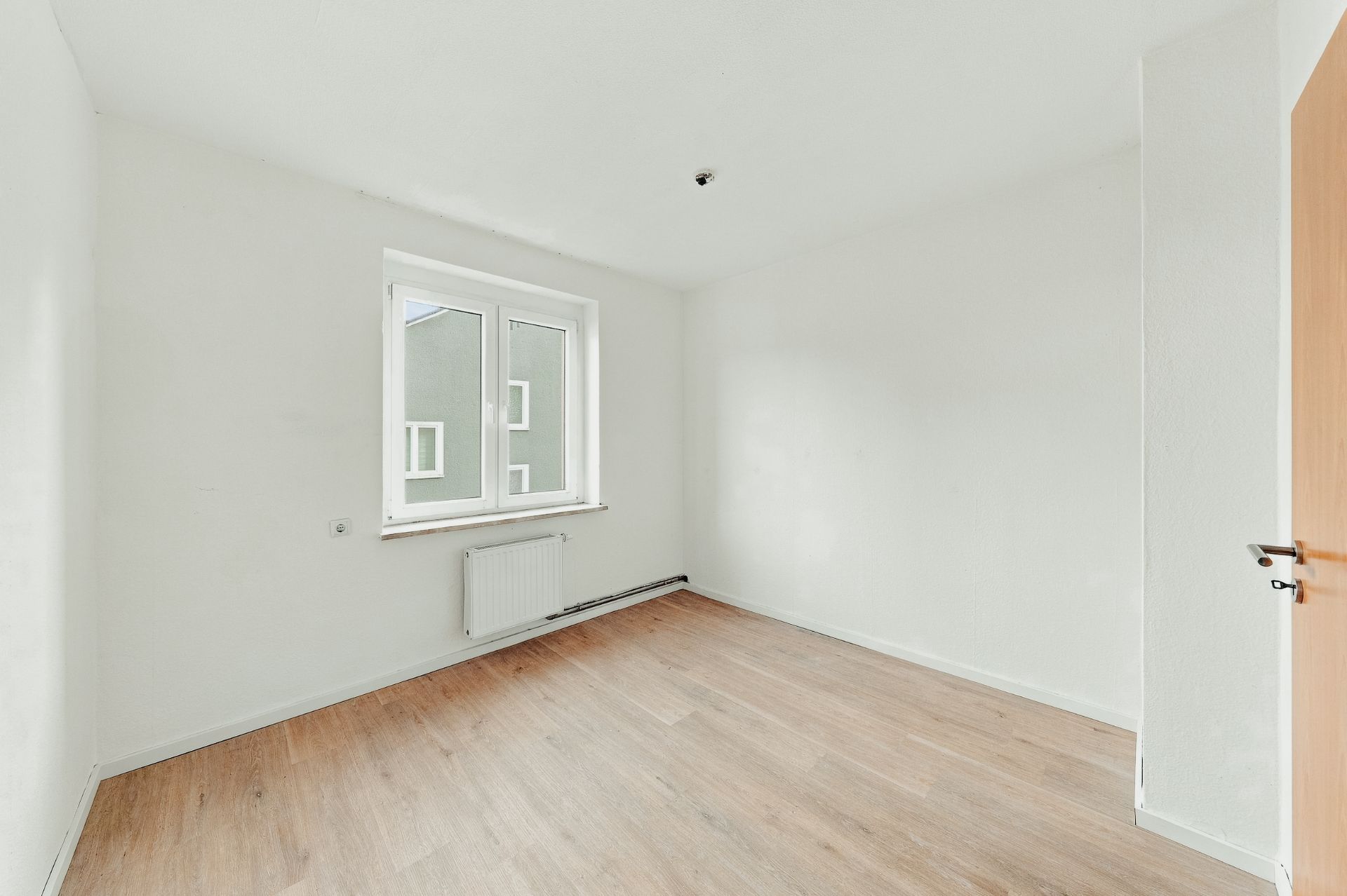 Helles, leeres Zimmer mit weißem Wänden, Fenster, Heizkörper und Holzboden.