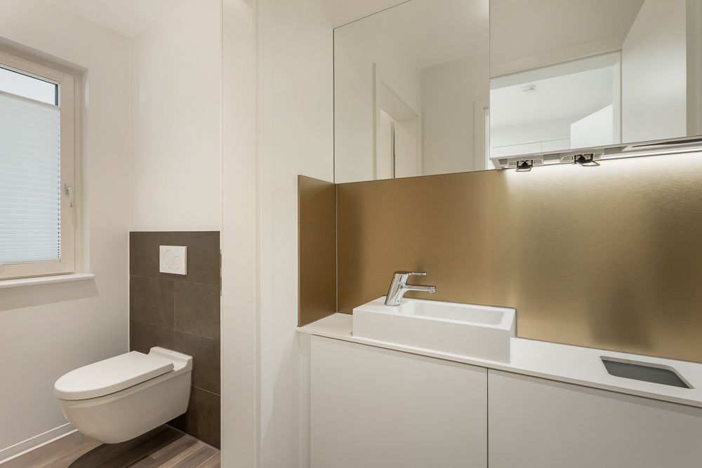 Modernes Badezimmer mit Wandtoilette und rechteckigem Waschbecken vor goldenem Spiegel.