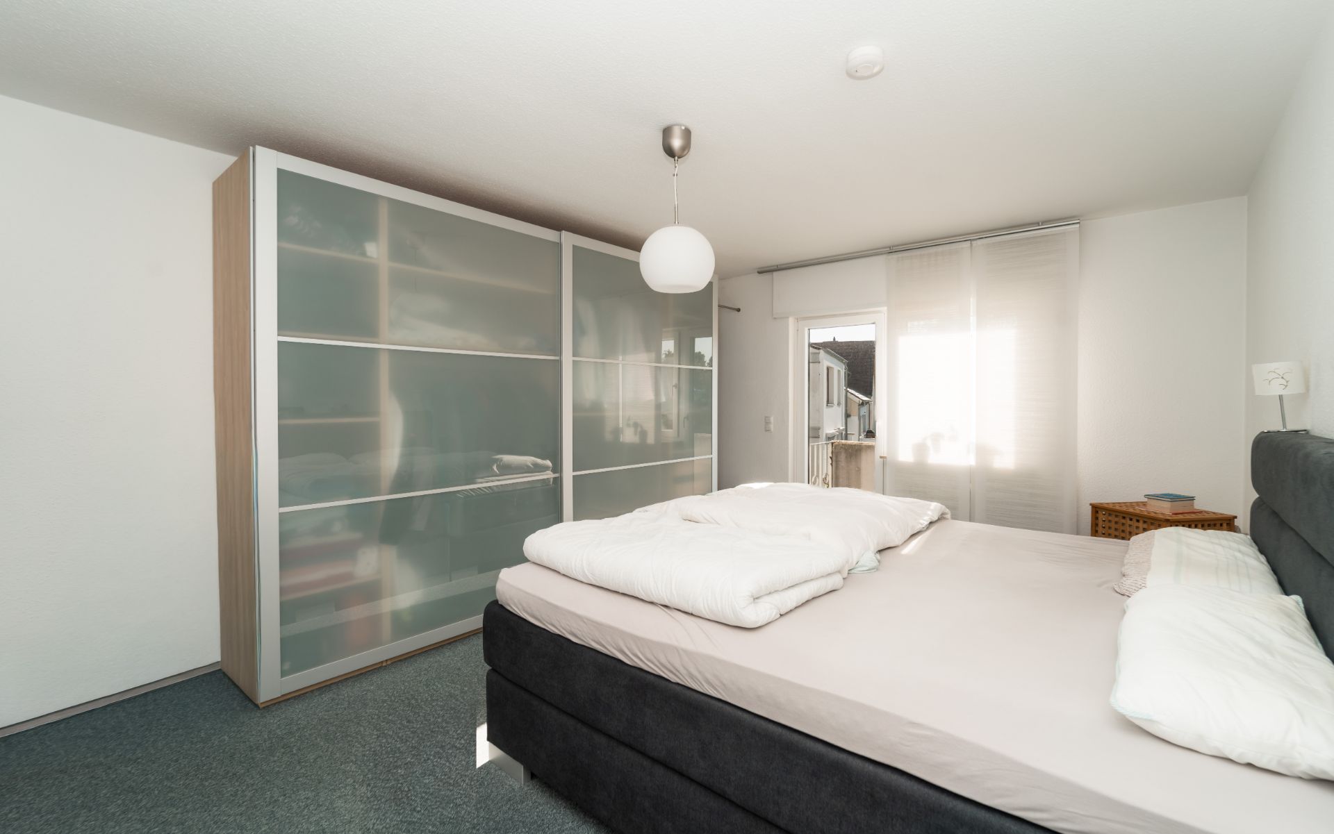 Schlafzimmer mit Doppelbett, großem Schrank mit Schiebetüren und Fenster mit Vorhang.