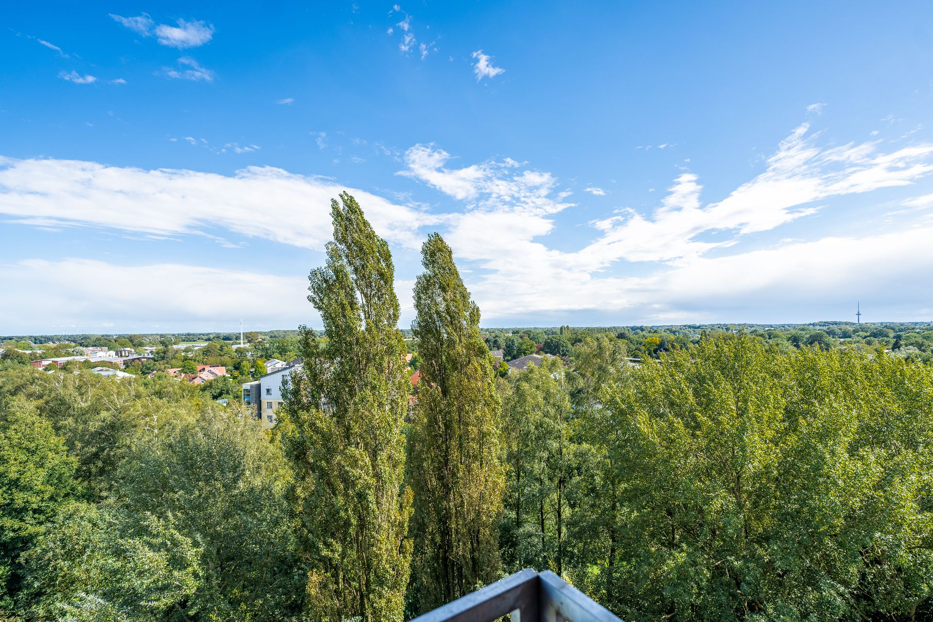 Blick von einem Balkon auf grüne Bäume und Wohngebäude unter blauem Himmel.