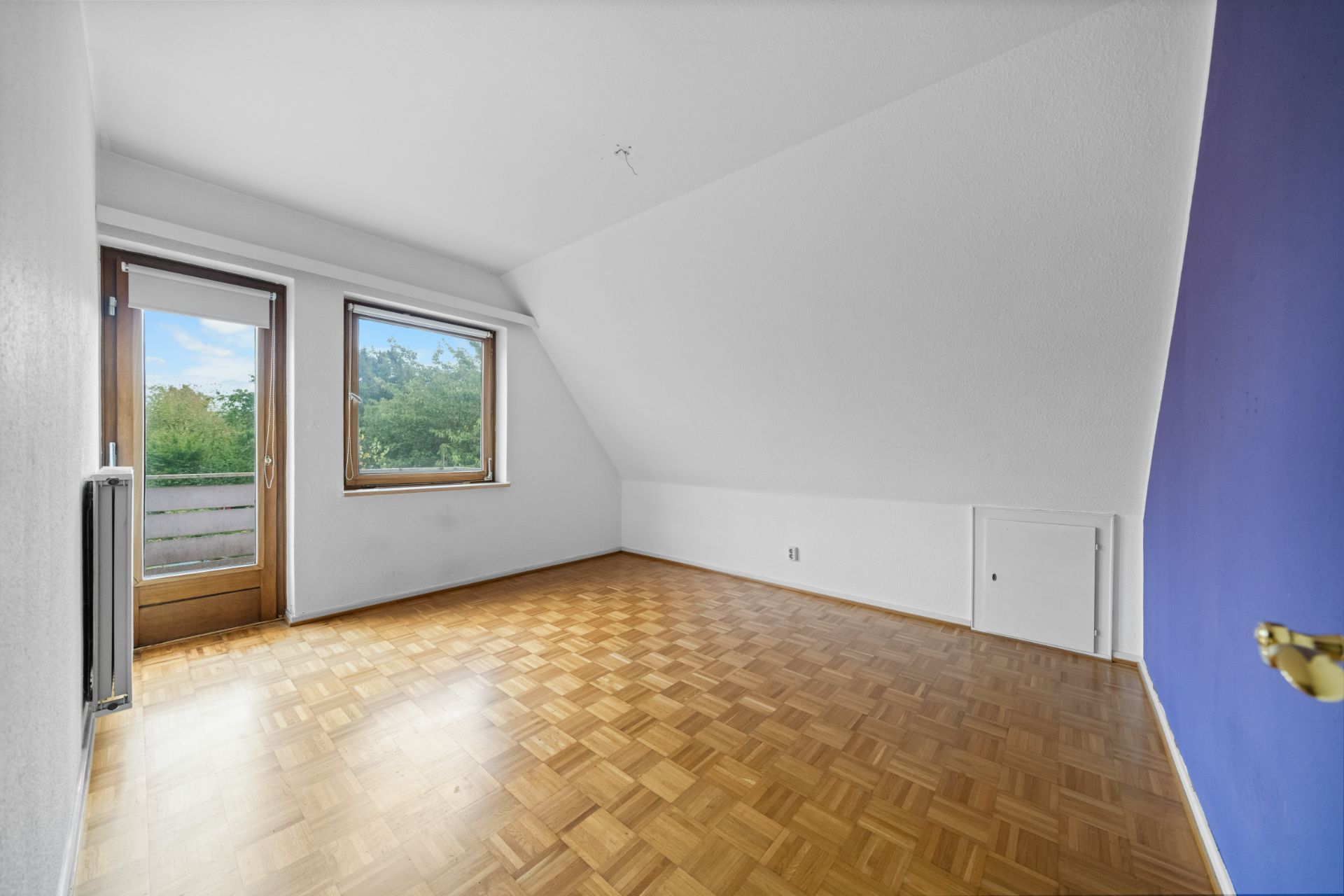 Leeres Zimmer mit Holzboden, weißer Wand, schräger Decke, Fenster und Tür zum Balkon.