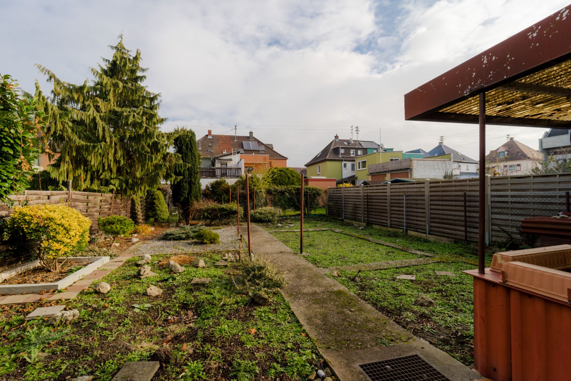 Garten mit Weg, Wiese, Bäumen, Sträuchern und Zaun neben Wohnhäusern bei bewölktem Wetter.