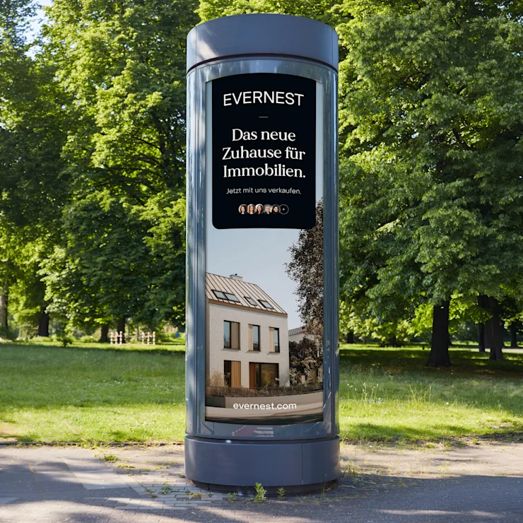 Werbesäule im Park mit Anzeige für Immobilien und Bild eines modernen Wohnhauses.