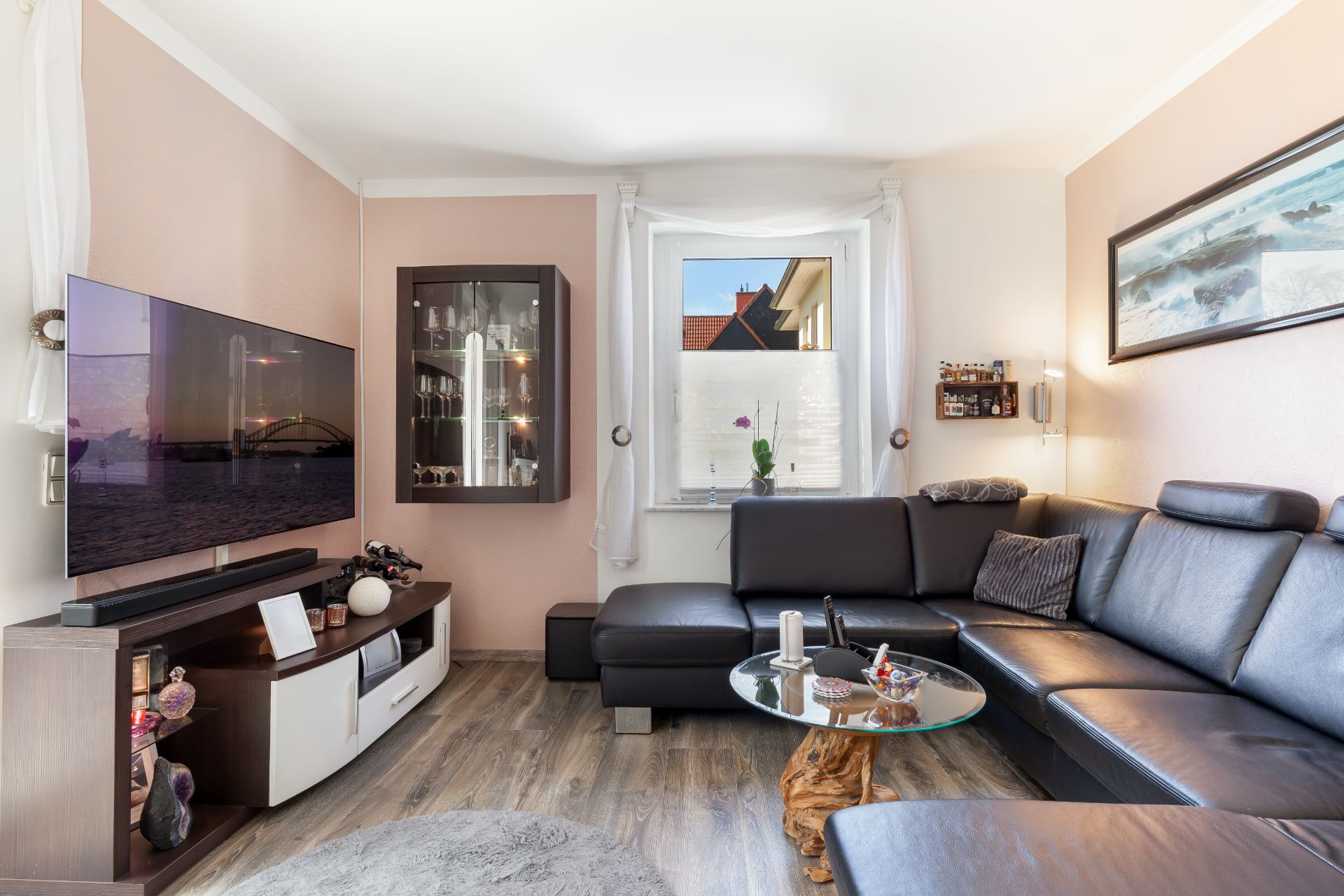 Modernes Wohnzimmer mit schwarzem Ecksofa, Wandfernseher und rundem Glastisch vor einem Fenster.