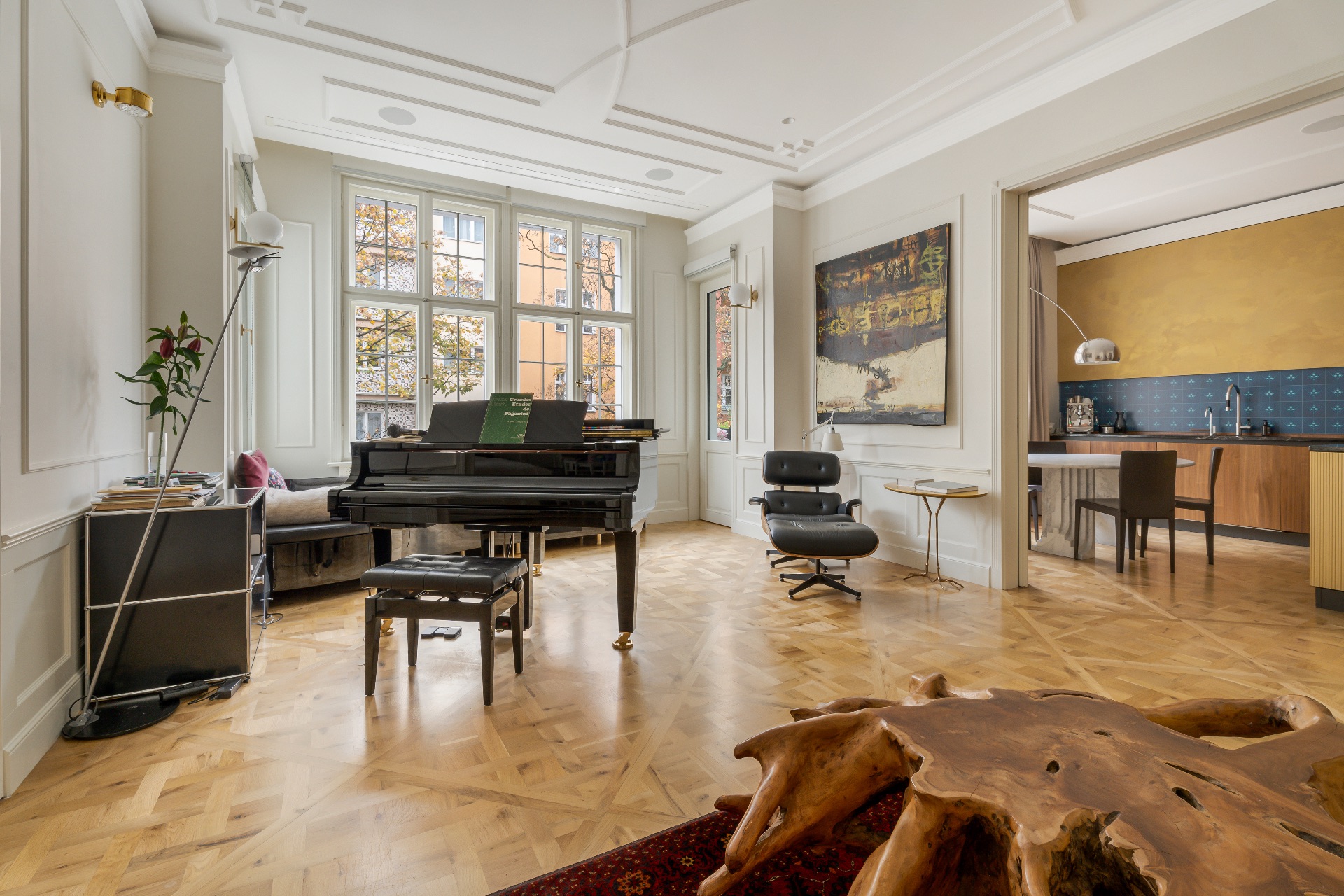 Helles Wohnzimmer mit Flügel, Sessel, großem Fenster und Blick in die offene Küche.