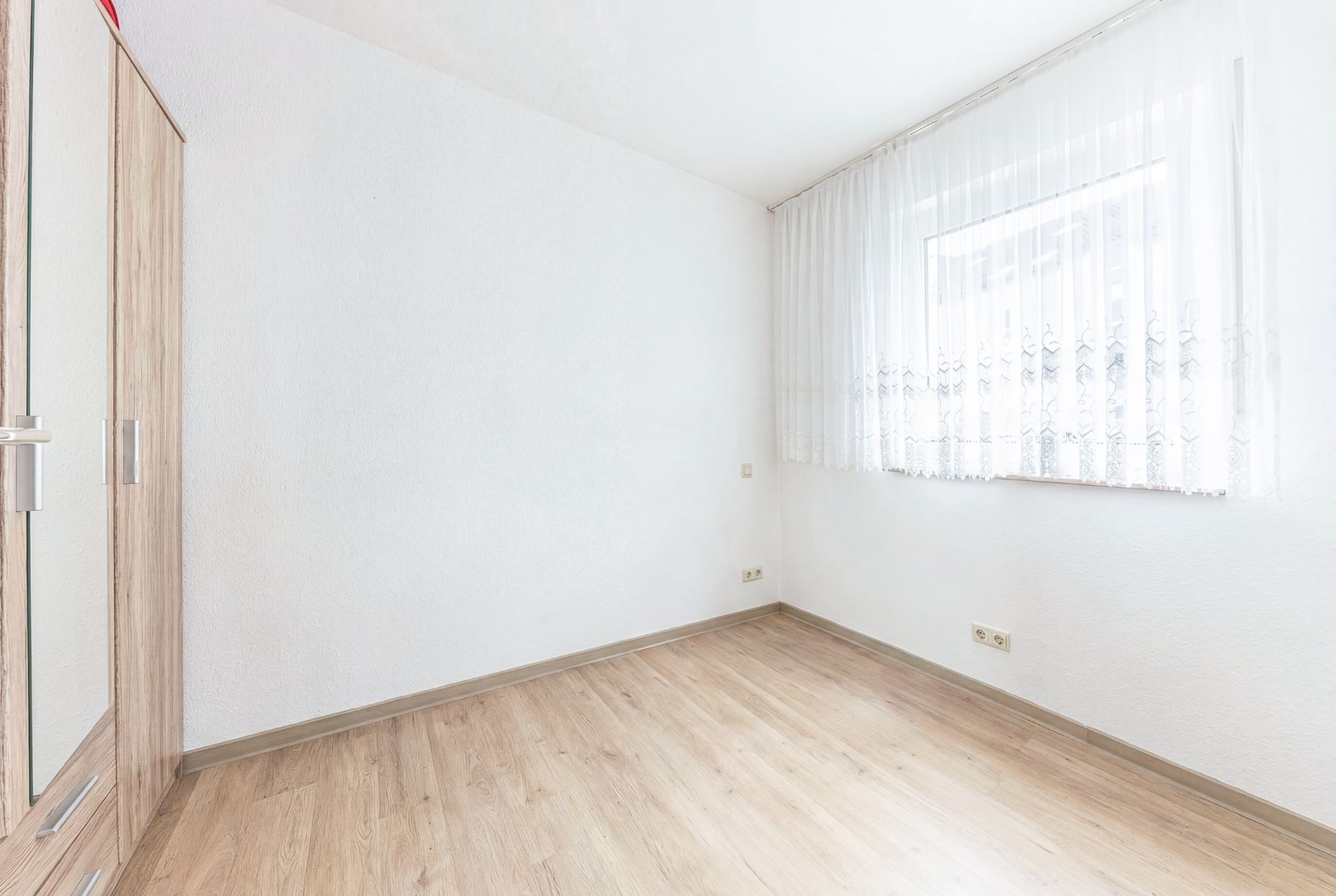 Leeres, helles Zimmer mit Holzfußboden, Fenster und Kleiderschrank an der linken Wand.