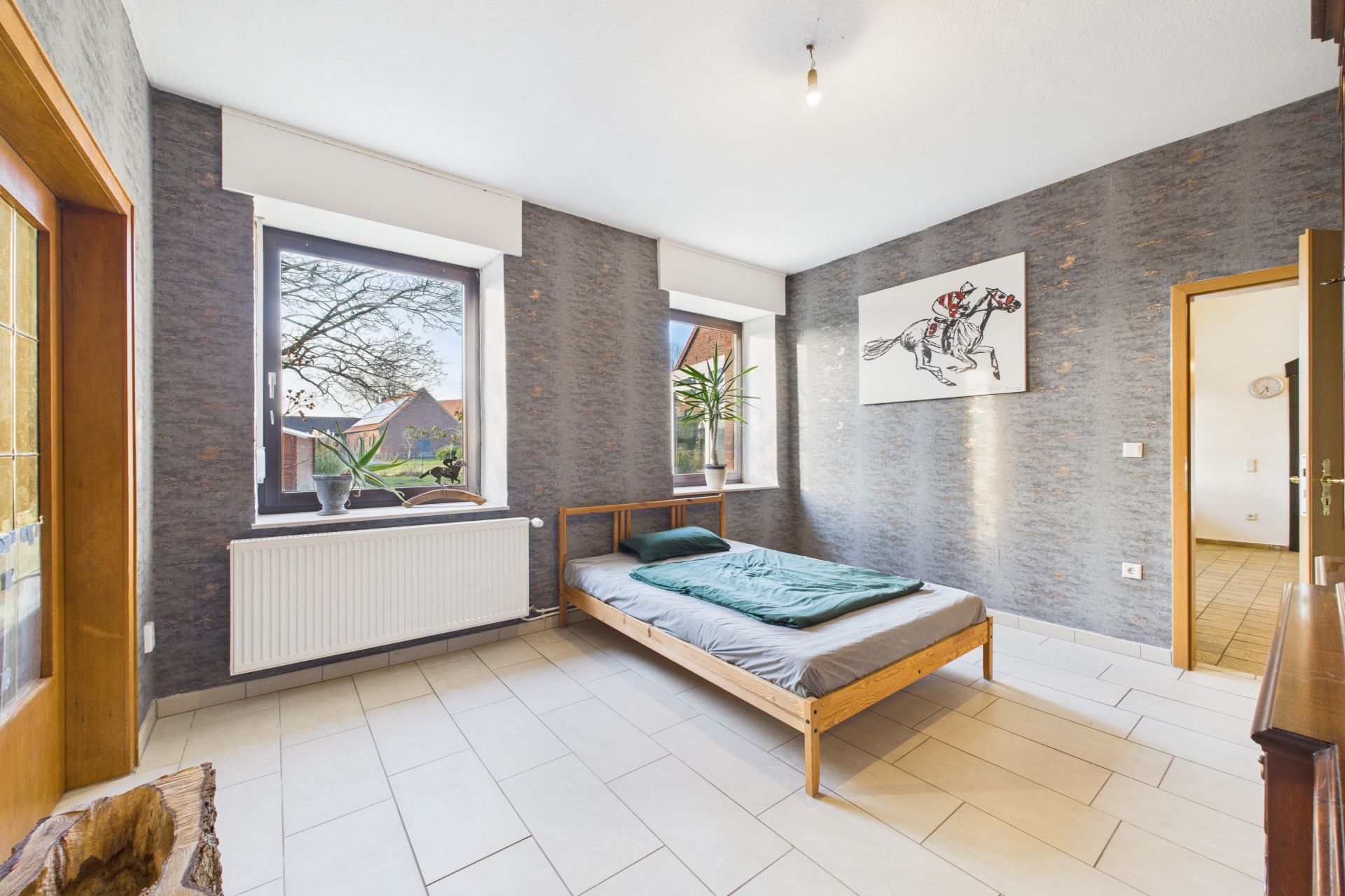 Helles Schlafzimmer mit Bett, zwei Fenstern, Pflanzen und Bild an der Wand.