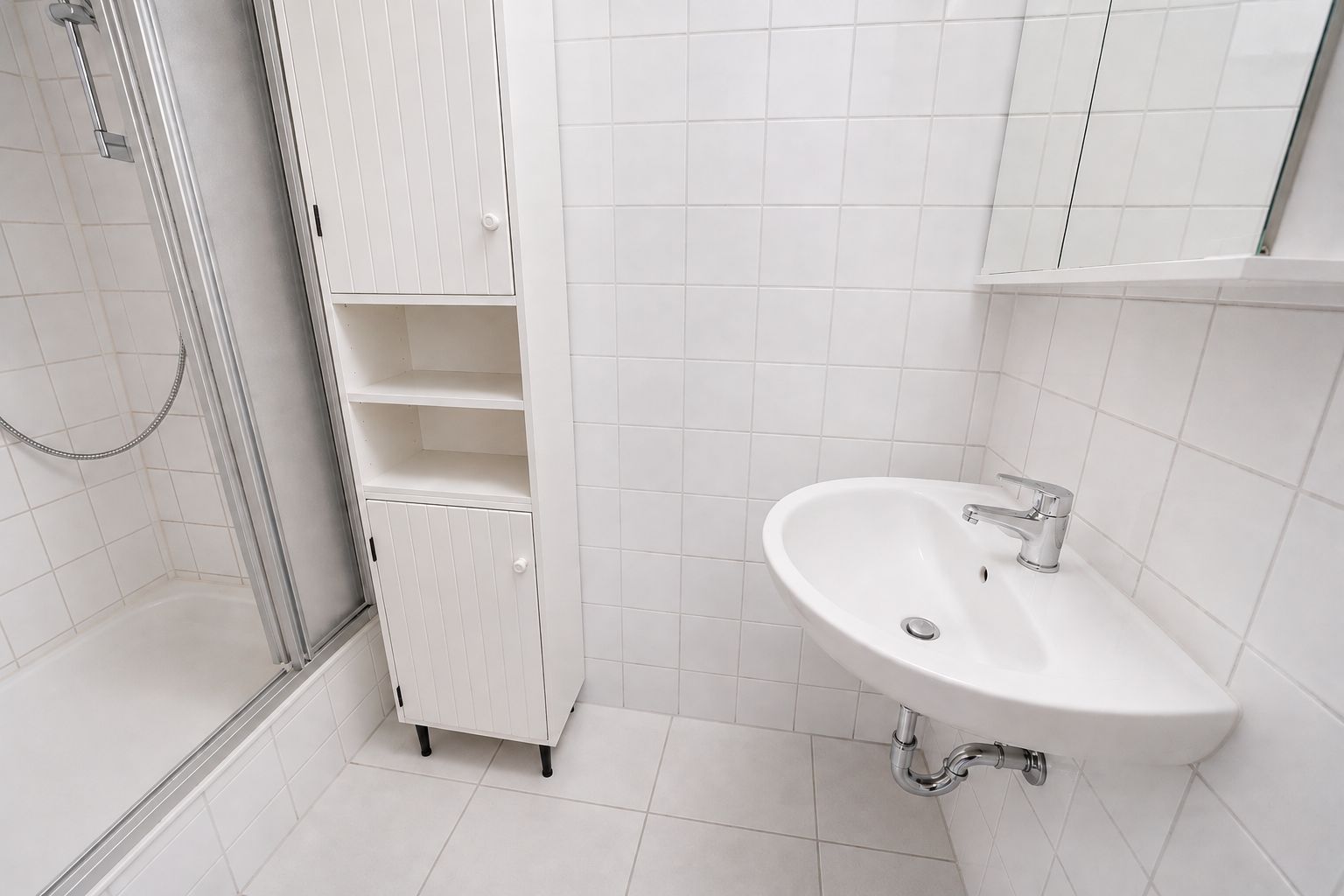 Helles Badezimmer mit Dusche, weißem Waschbecken und weißem Schrank auf weißen Fliesen.