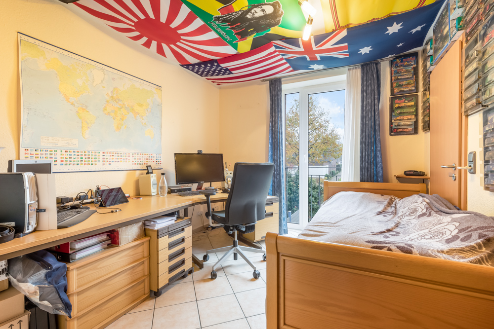 Arbeitszimmer mit Schreibtisch, Stuhl, Computer, Weltkarte und Einzelbett, Flaggen an der Decke.