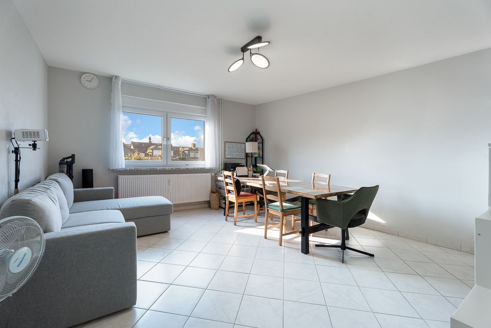 Helles Wohnzimmer mit grauem Sofa, Esstisch und fünf Stühlen auf weiß gefliestem Boden.