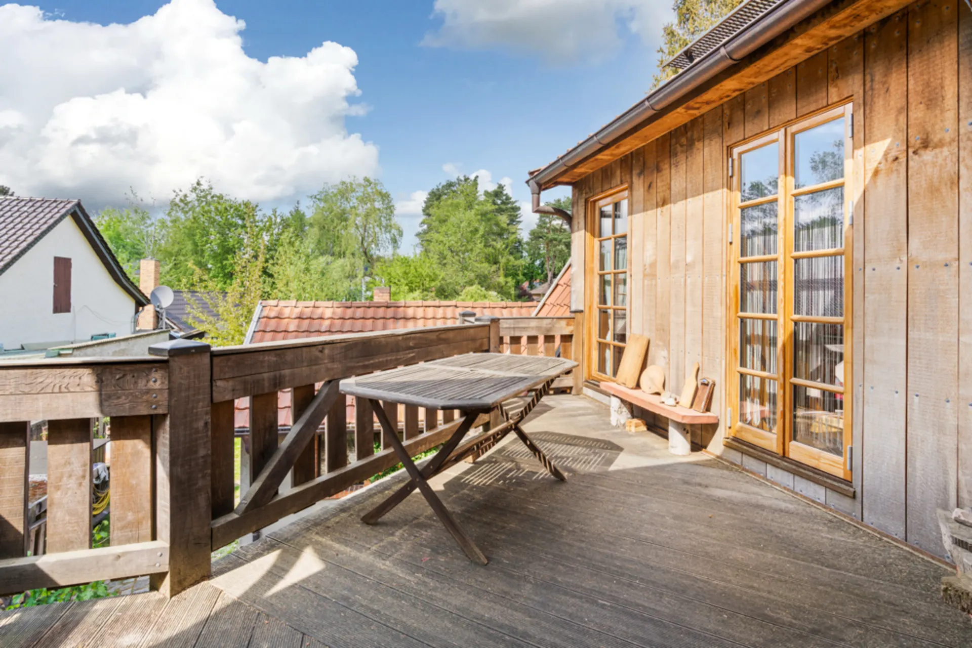 Holzbalkon mit Tisch und Bank, Holzwand und großen Fenstern, Blick auf Bäume und Nachbarhäuser.