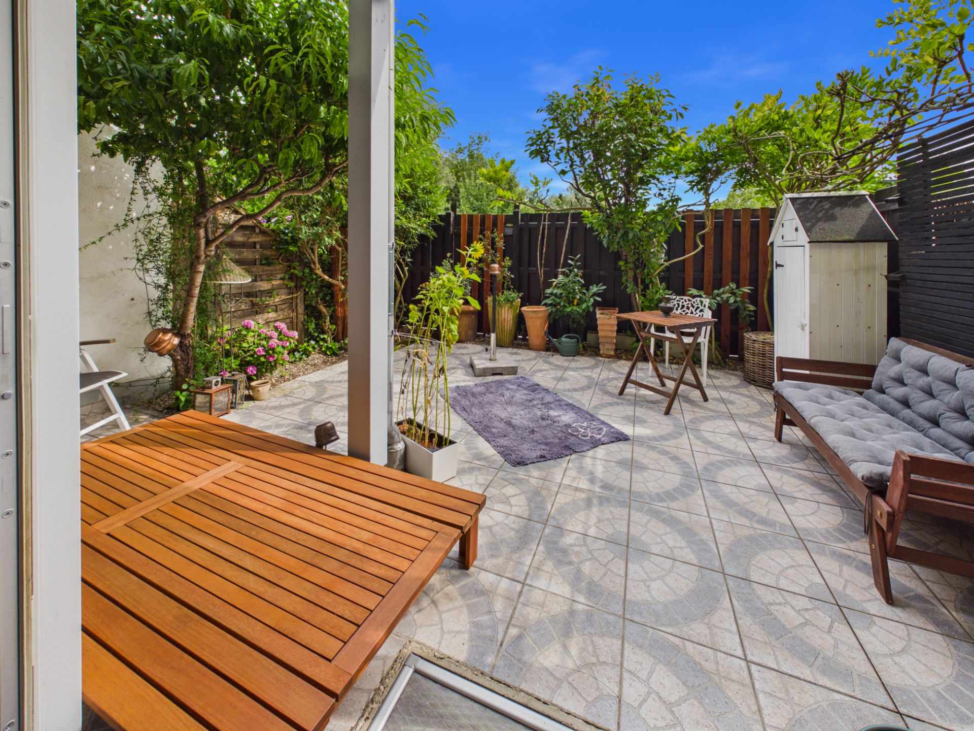Geflieste Terrasse mit Holzmöbeln, Pflanzen, Sitzbank und kleinem Tisch im Garten.