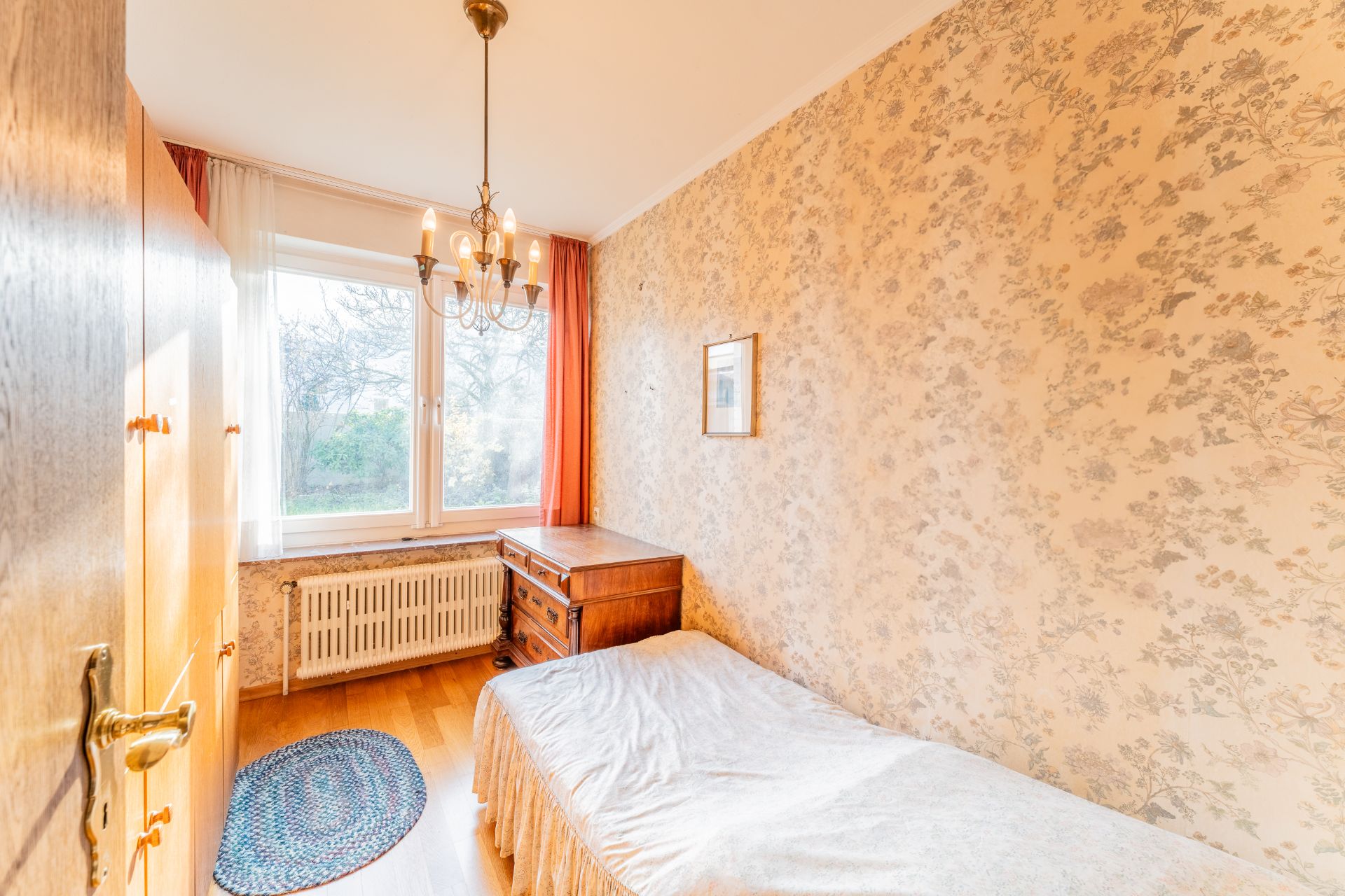 Kleines Schlafzimmer mit Einzelbett, Holzschrank, Schreibtisch und Fenster mit Vorhang.