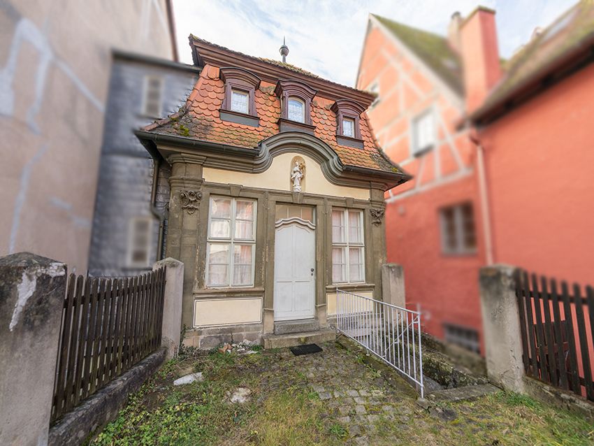 Kleines, zweistöckiges Haus mit rotem Dach und weißen Fenstern, kleiner Vorgarten, Zaun links und rechts.