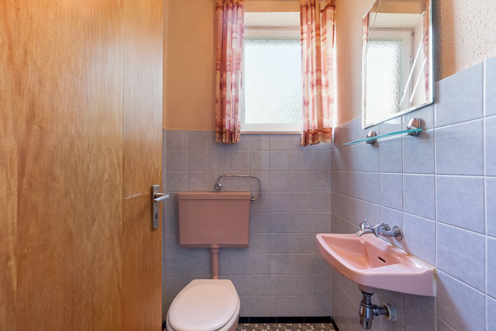 Kleines Badezimmer mit Toilette, rosa Waschbecken, Spiegel und Fenster mit Vorhängen.