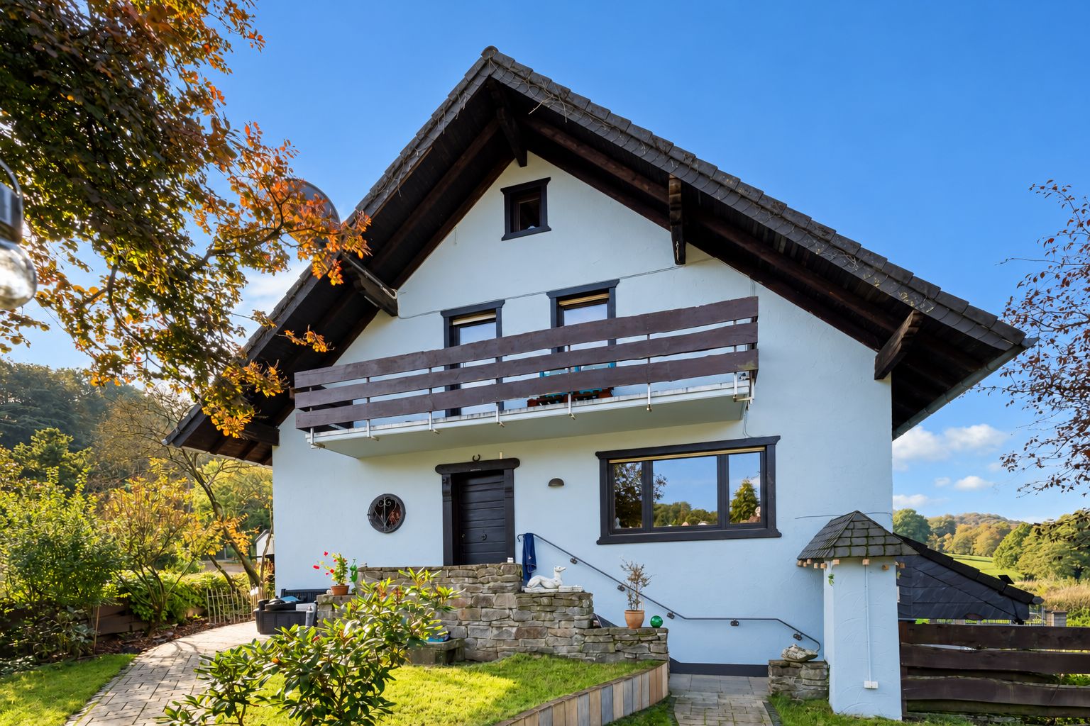 Zweistöckiges weißes Wohnhaus mit Balkon, Garten und Steintreppe vor blauem Himmel.