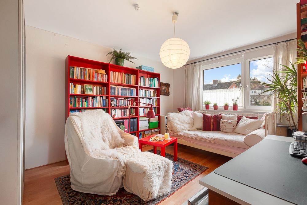 Helles Wohnzimmer mit rotem Bücherregal, Sofa, Sessel, Teppich und großem Fenster.