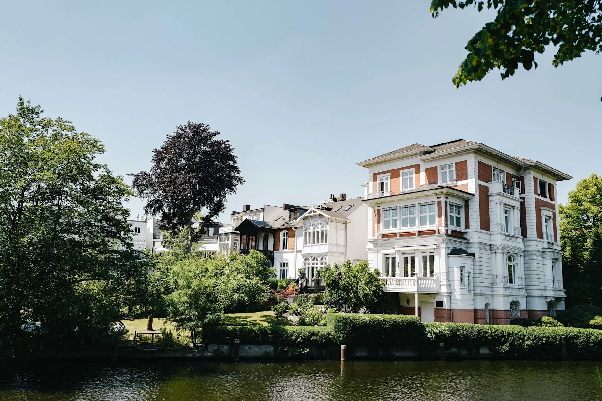 Großes Wohnhaus mit Garten am Wasser, umgeben von Bäumen bei klarem Himmel.