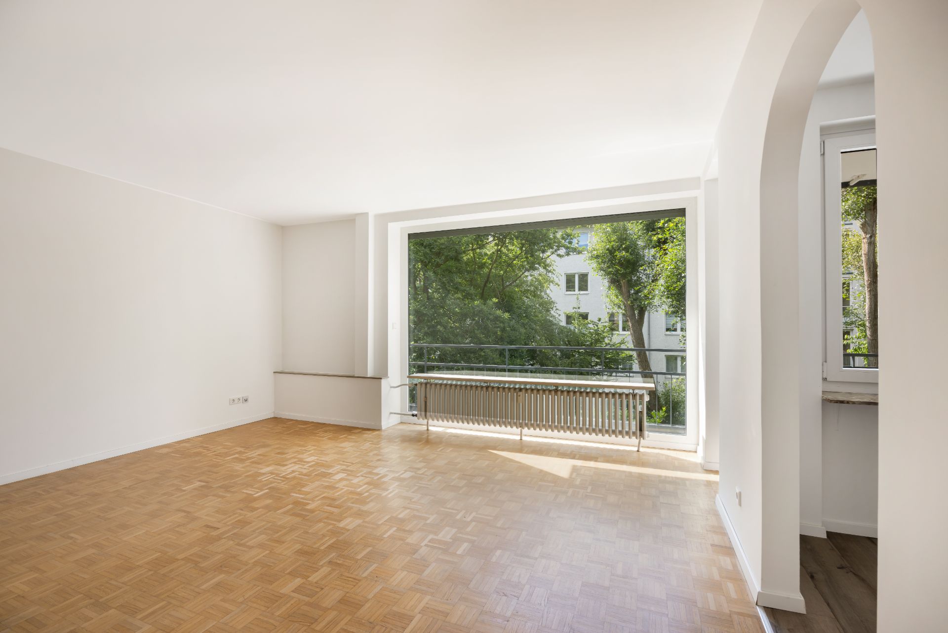 Helles Wohnzimmer mit Parkettboden und großem Fenster mit Blick ins Grüne.
