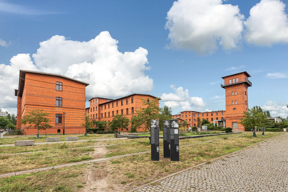Mehrere rote Backsteingebäude mit Bäumen und Weg auf einem Grundstück unter blauem Himmel.