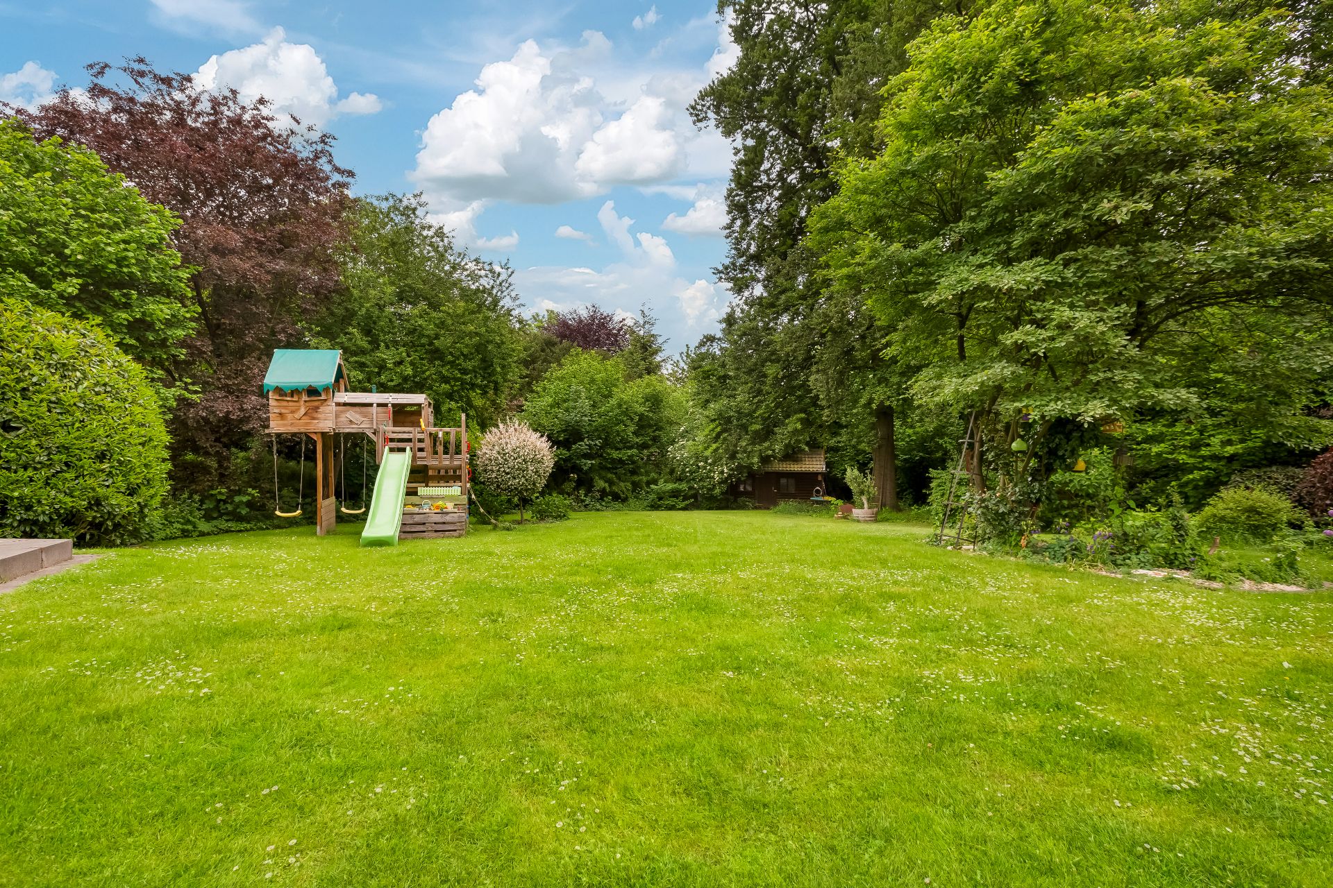 Großer grüner Garten mit Bäumen, Rutsche, Schaukel und Holzhäuschen im Hintergrund.