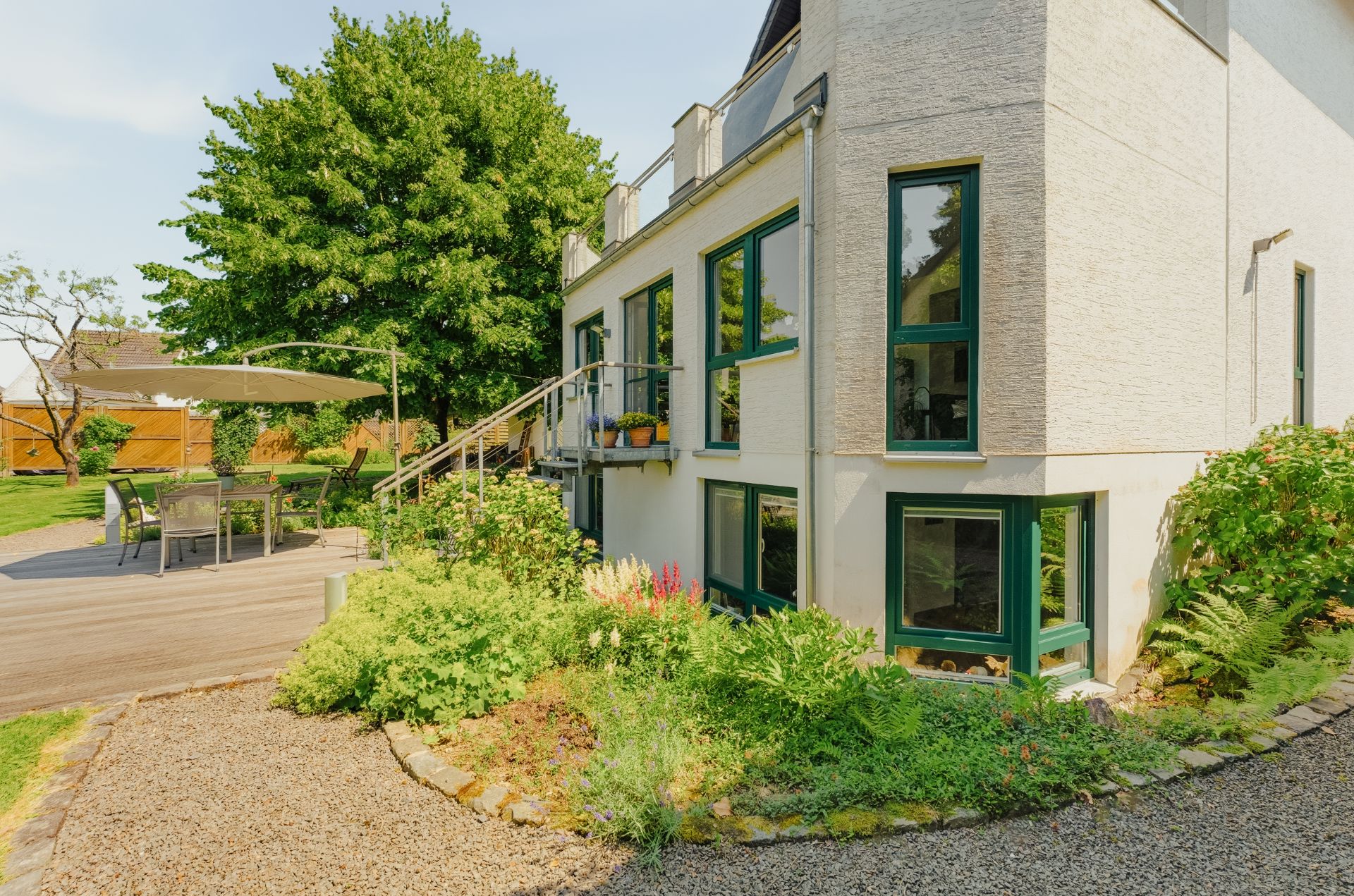 Haus mit Garten, Terrasse mit Tisch und Stühlen, umliegende Pflanzen und Baum.