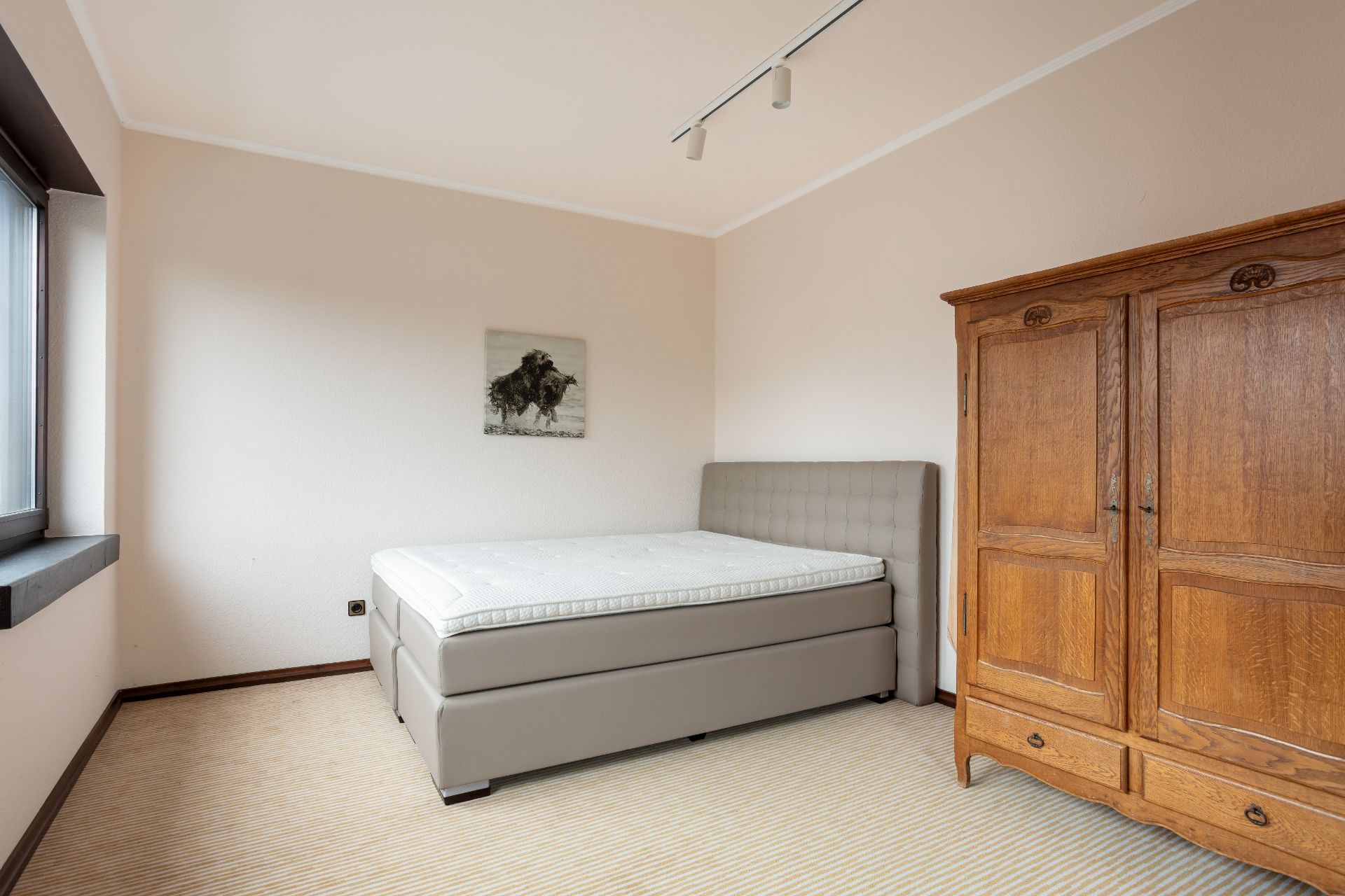 Schlafzimmer mit grauem Bett, Holzschrank und Fenster, hell gestrichene Wände.