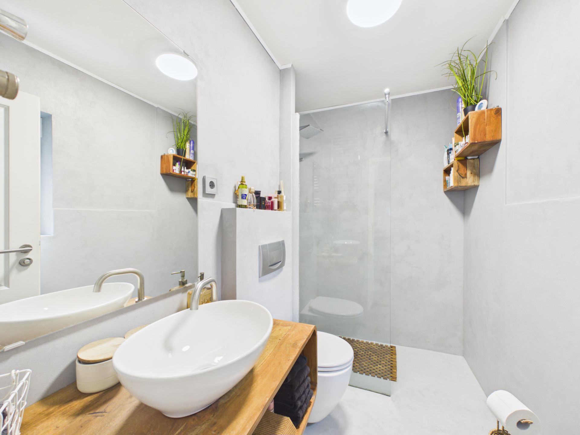 Modernes Badezimmer mit Waschtisch, Spiegel, WC und begehbarer Dusche, Regale mit Pflanzen und Pflegeprodukten.