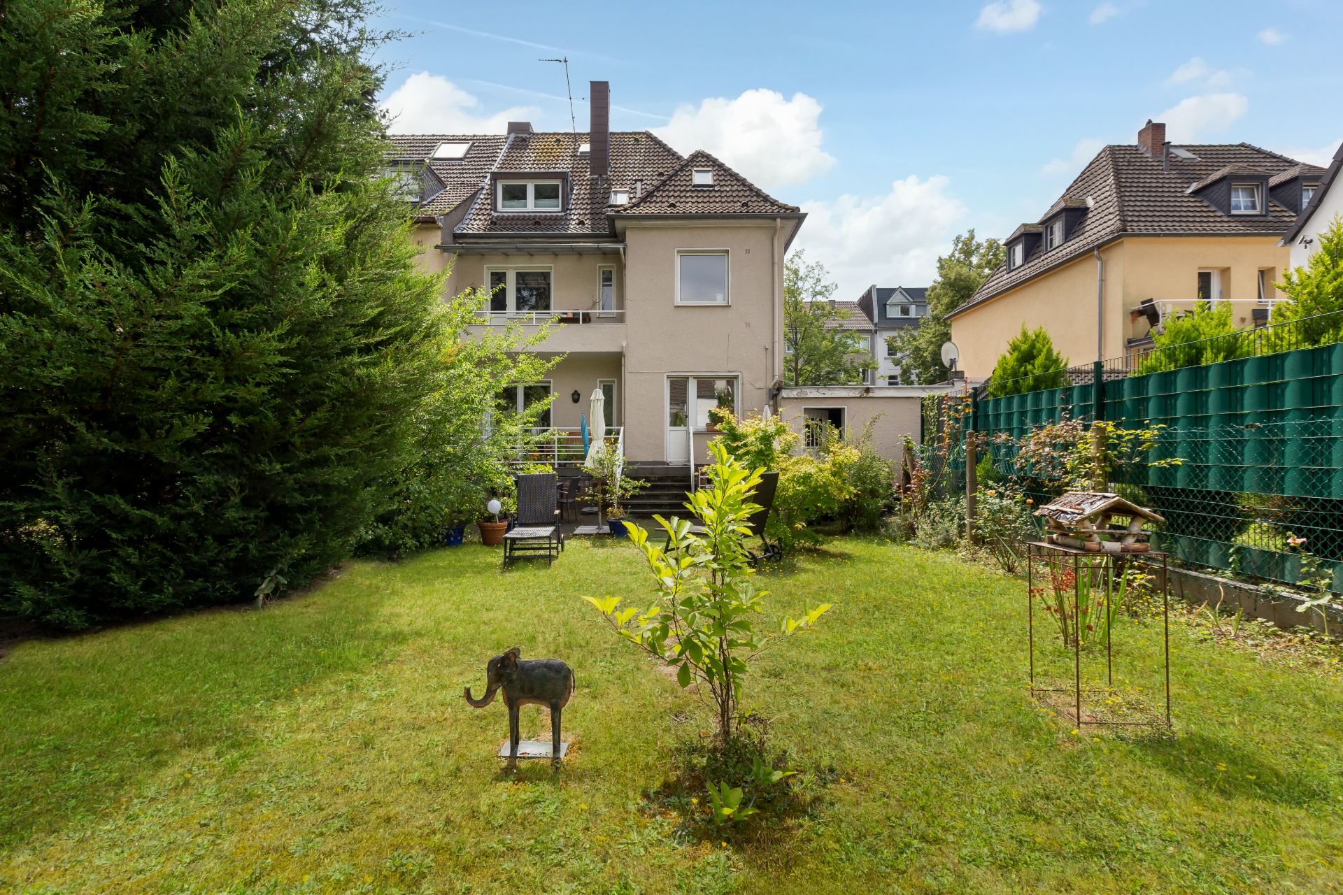 Grüner Garten mit kleiner Elefantenfigur, Gartenmöbeln und Bäumen hinter einem Wohnhaus.