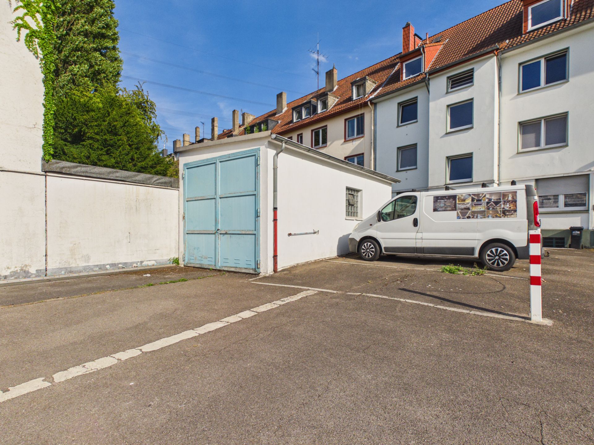 Hof mit kleinem Garagengebäude, weißem Transporter und Wohnhäusern im Hintergrund.