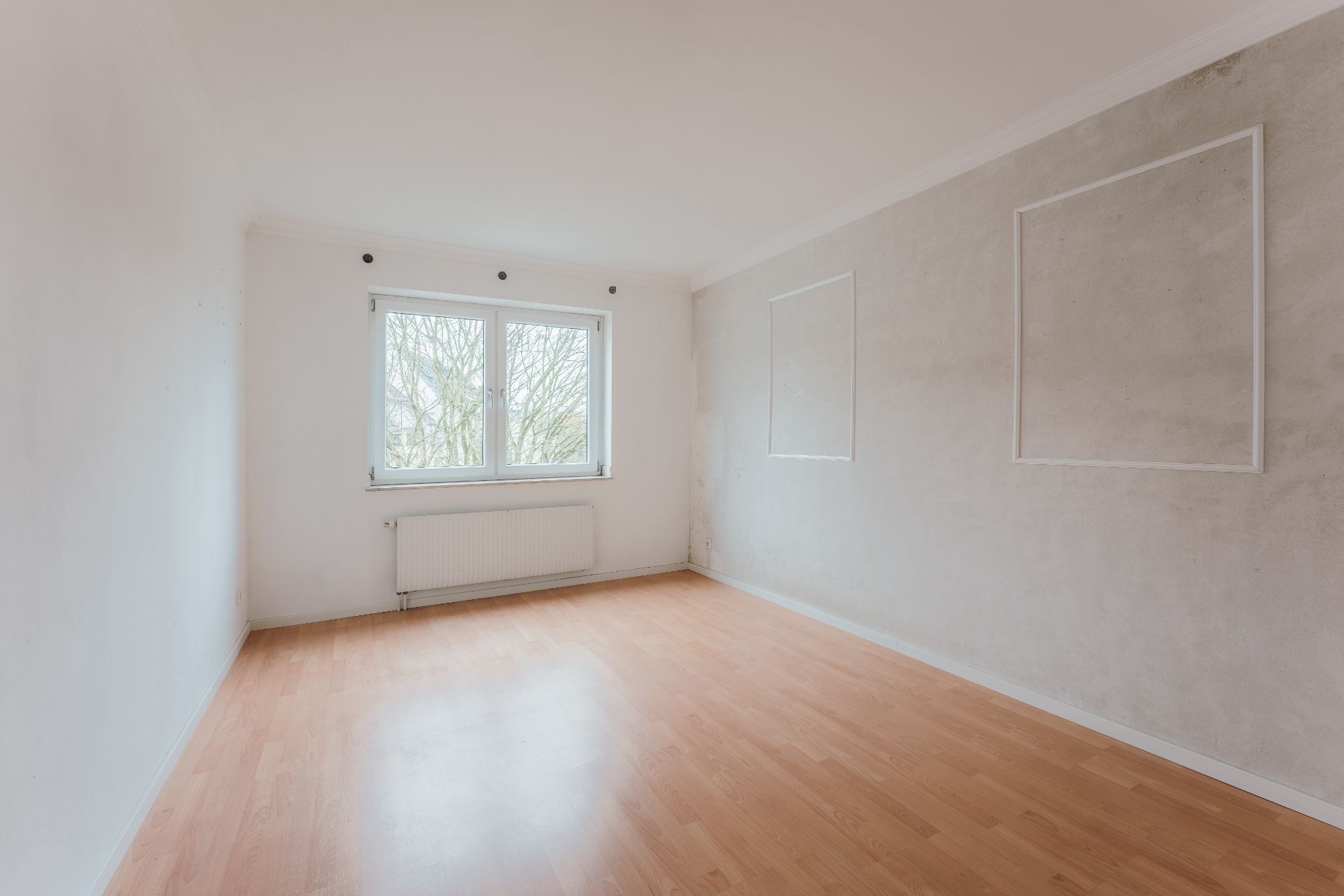 Leerer Raum mit hellem Laminatboden, weißer Wand, Fenster und Heizkörper.