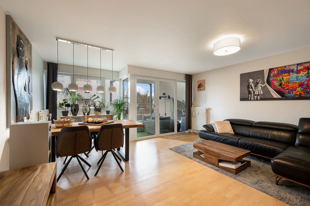 Helles Wohnzimmer mit schwarzem Sofa, Esstisch, moderner Beleuchtung und Zugang zum Balkon.