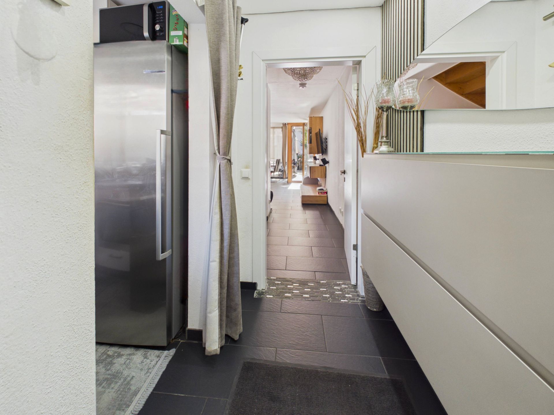 Flur mit schwarzem Boden, großem Kühlschrank, Spiegel und Blick ins Wohnzimmer.