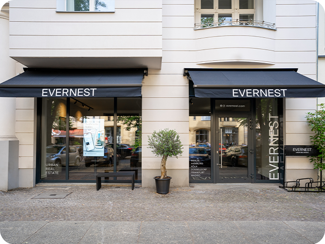 Schaufenster und Eingang eines Geschäfts mit zwei schwarzen Markisen und einem kleinen Baum im Topf davor.