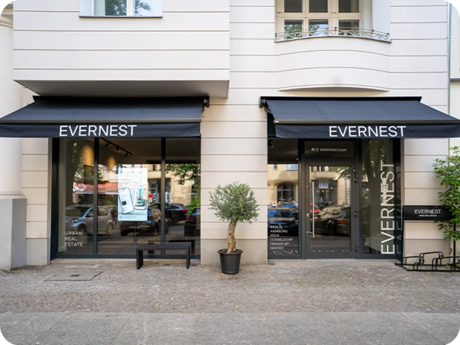 Schaufenster und Eingang eines Geschäfts mit zwei schwarzen Markisen und einem kleinen Baum im Topf davor.