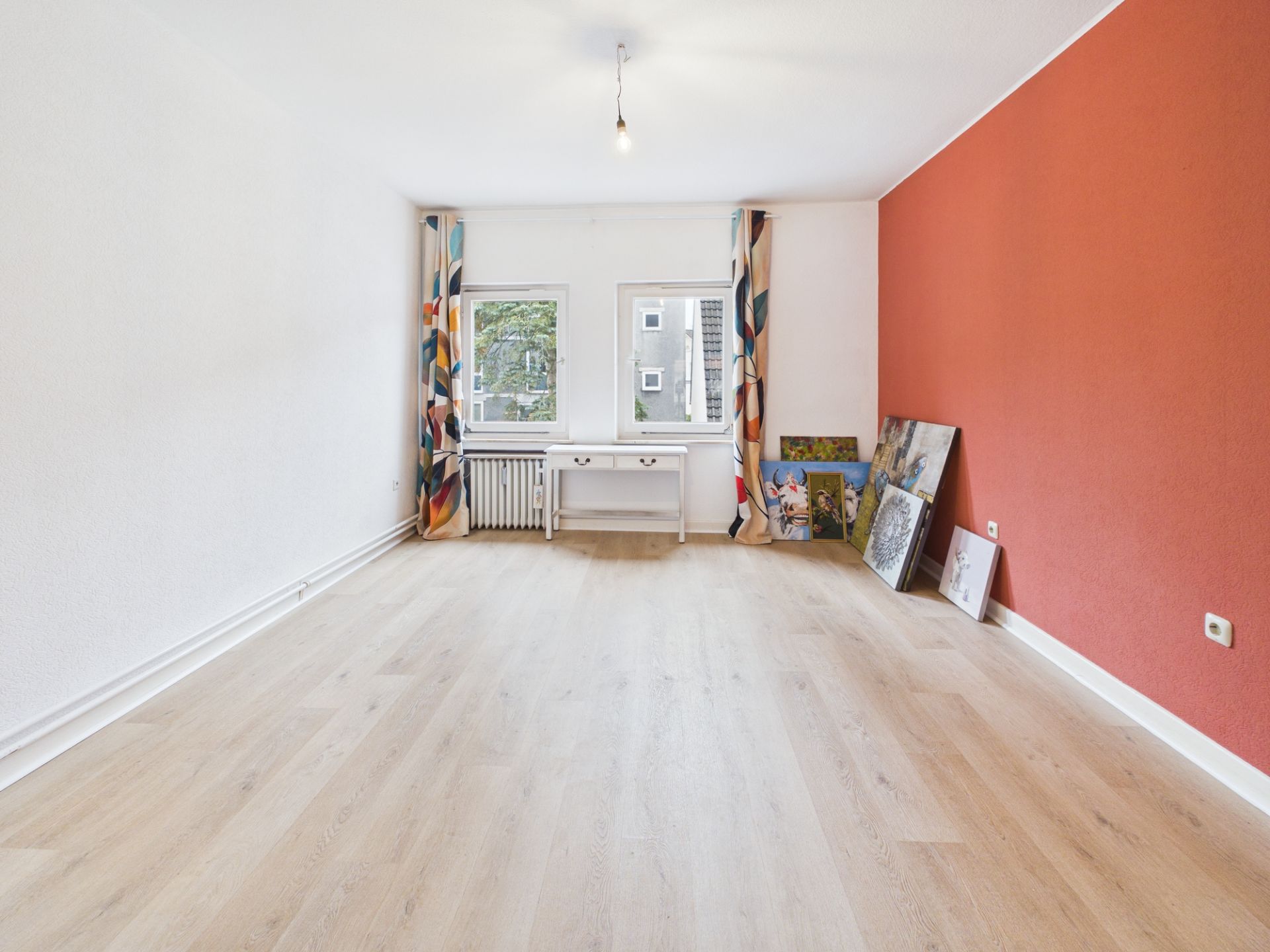 Helles Zimmer mit Holzboden, roter Wand, Schreibtisch bei zwei Fenstern und Bildern an der Wand.
