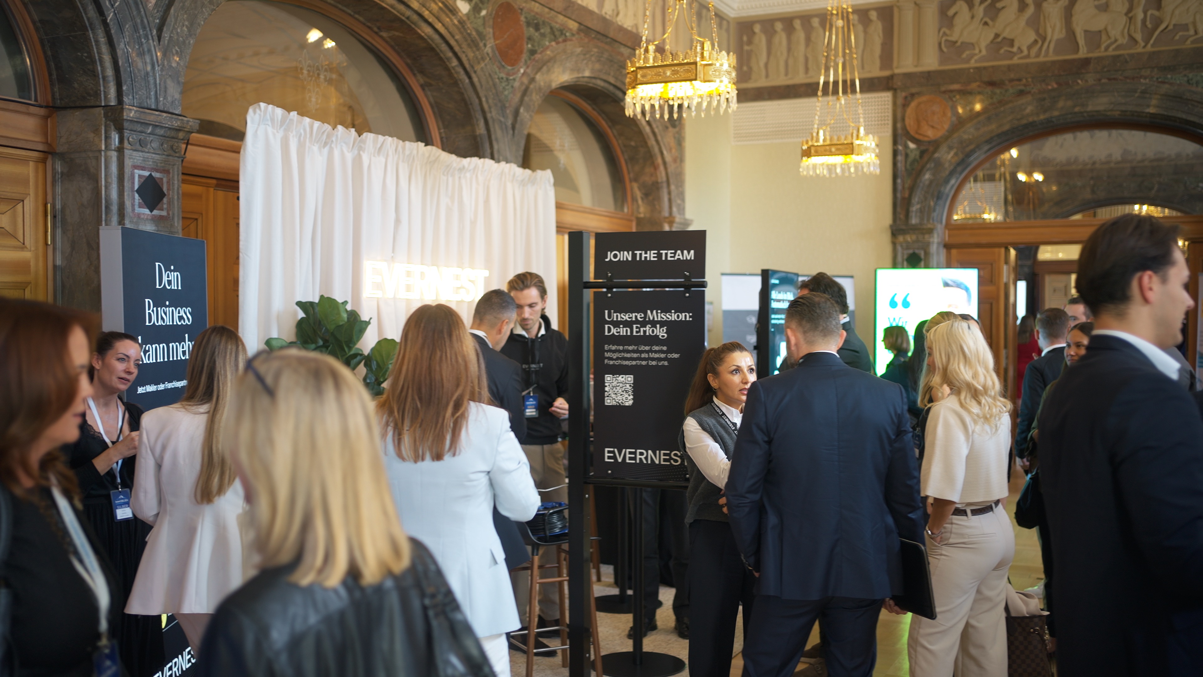 Mehrere Menschen im Gespräch in einem eleganten Foyer mit Marmorsäulen und Infotafeln.