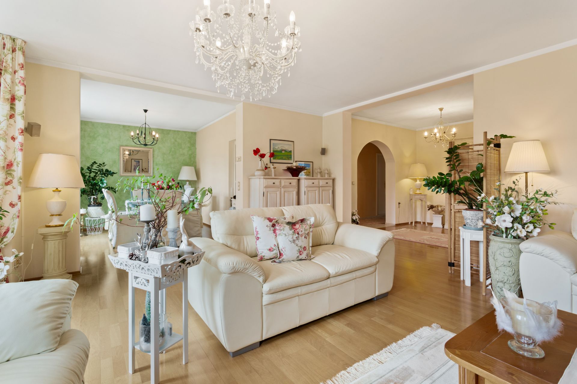 Helles Wohnzimmer mit cremefarbenen Sofas, Kronleuchter und vielen Pflanzen.