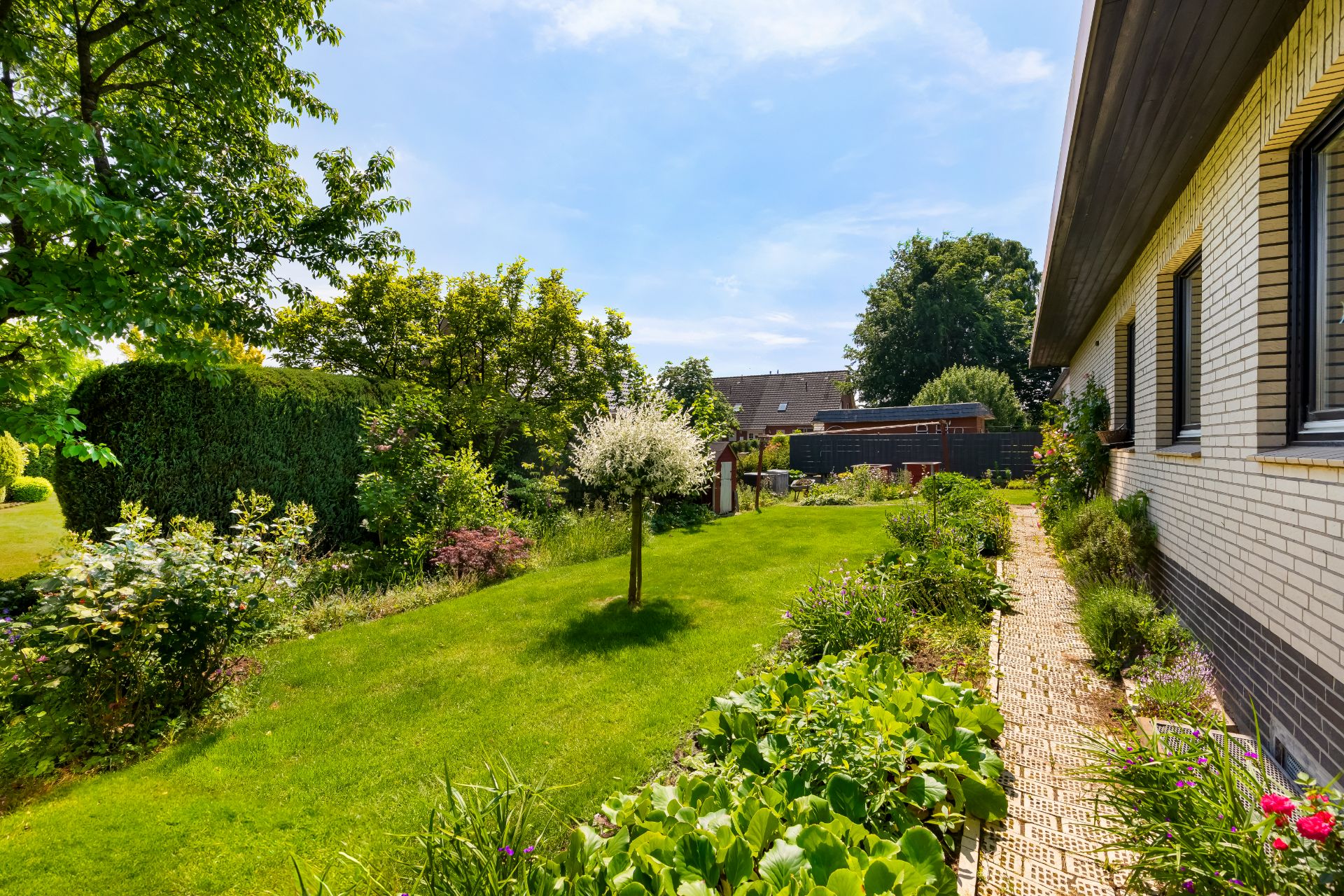 Gepflegter Garten mit Rasen, Bäumen, Sträuchern und Blumenbeeten neben einem Wohnhaus.