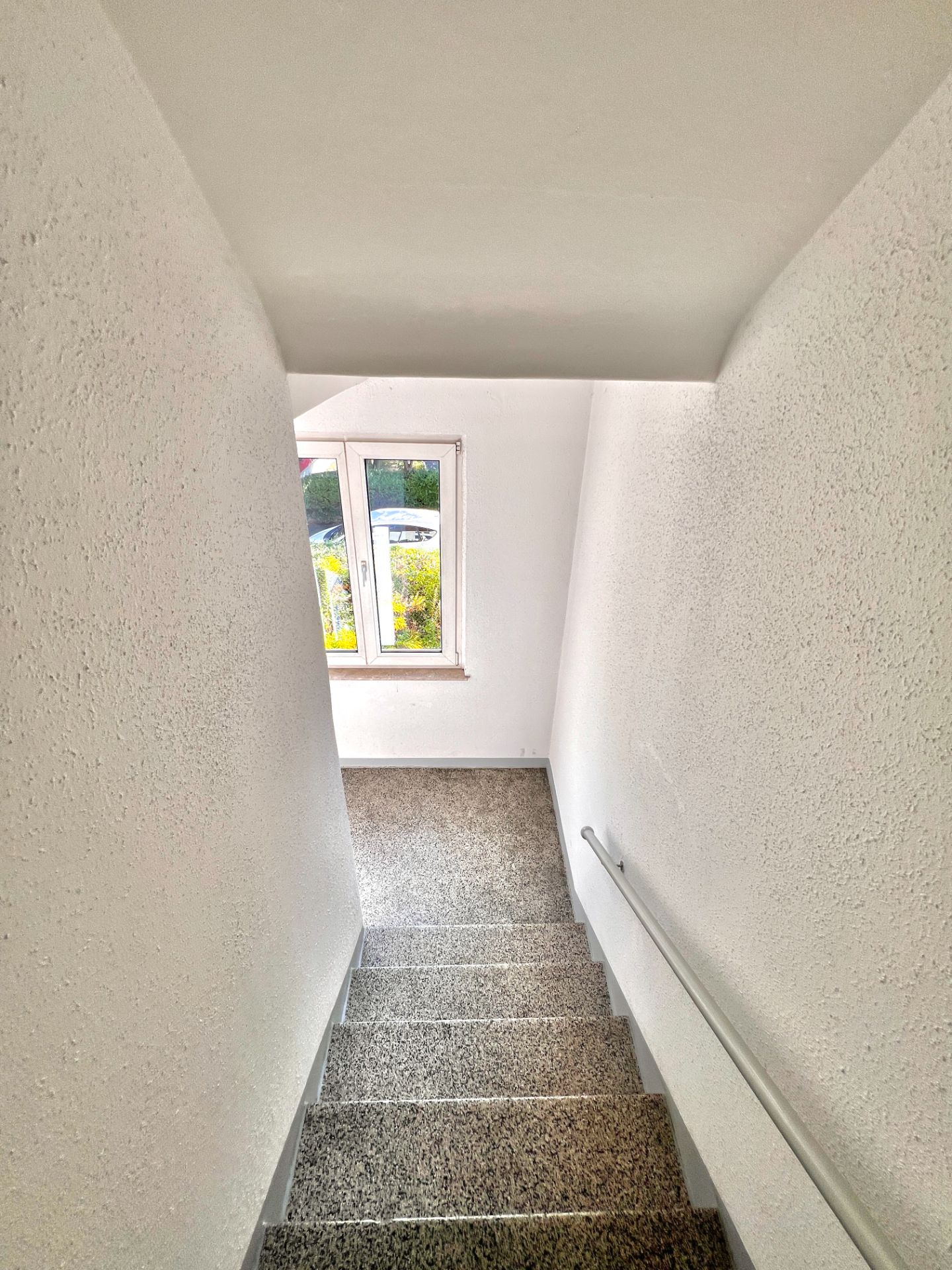 Treppe mit grauen Stufen und Handlauf, am Ende ein Fenster mit Blick nach draußen.