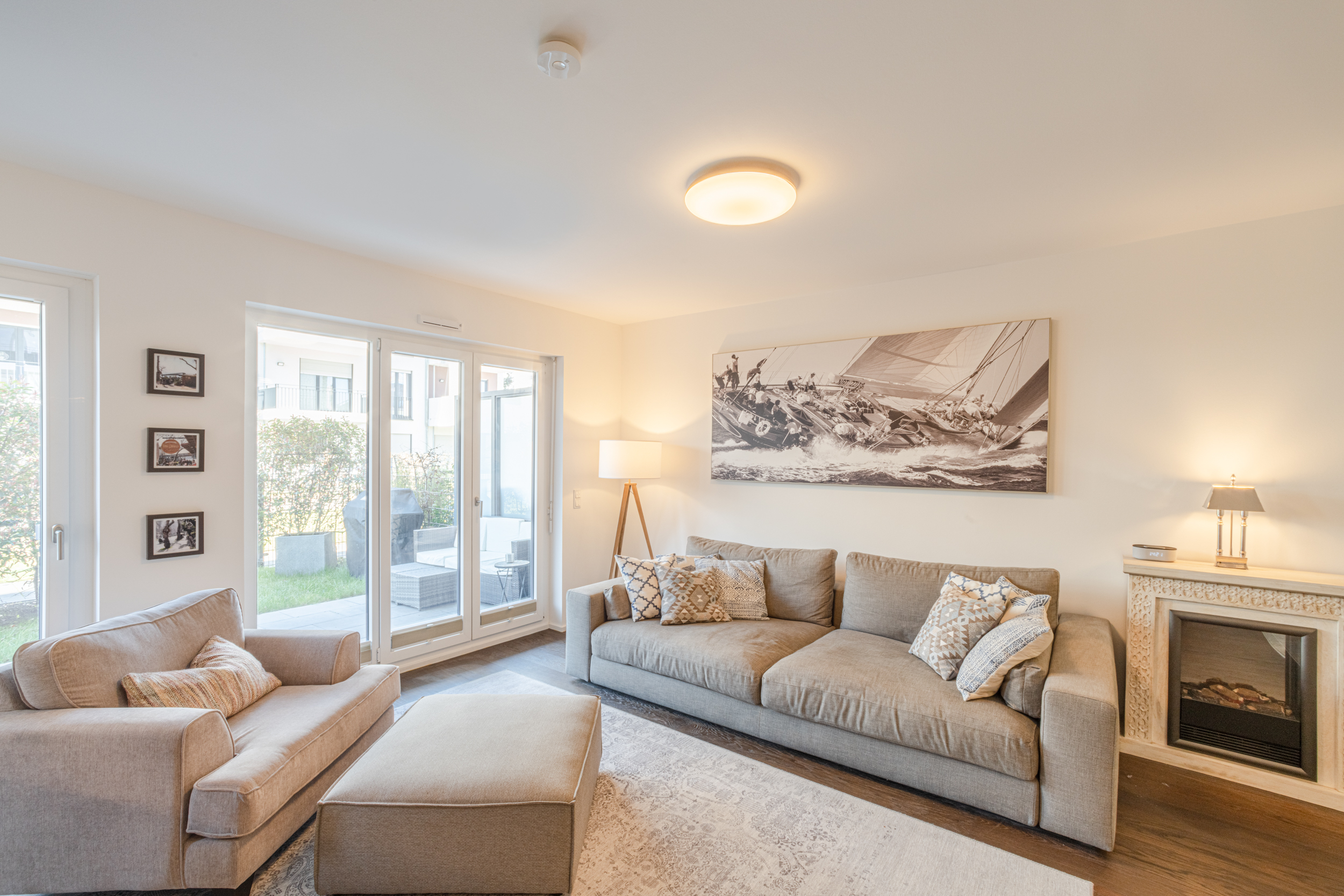 Helles Wohnzimmer mit Sofa, Sessel, Kamin, großem Fenster und Wandbild eines Segelbootes.