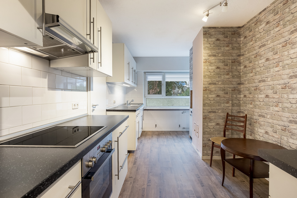 Moderne Einbauküche mit weißem Mobiliar, Holzfußboden und kleinem Esstisch mit Stuhl an der Backsteinwand.