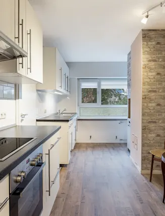 Moderne Einbauküche mit weißem Mobiliar, Holzfußboden und kleinem Esstisch mit Stuhl an der Backsteinwand.