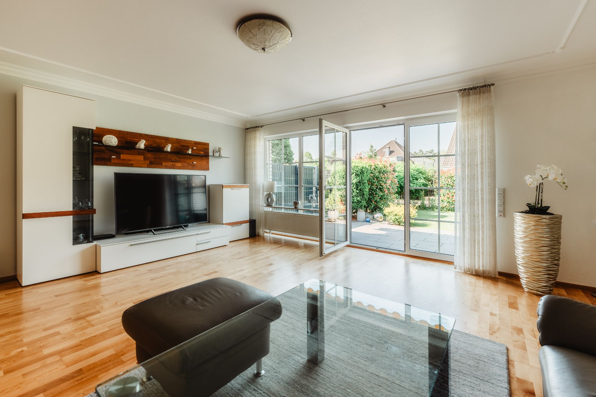 Helles Wohnzimmer mit Fernseher, Glas-Couchtisch und großen Fenstern mit Blick auf den Garten.