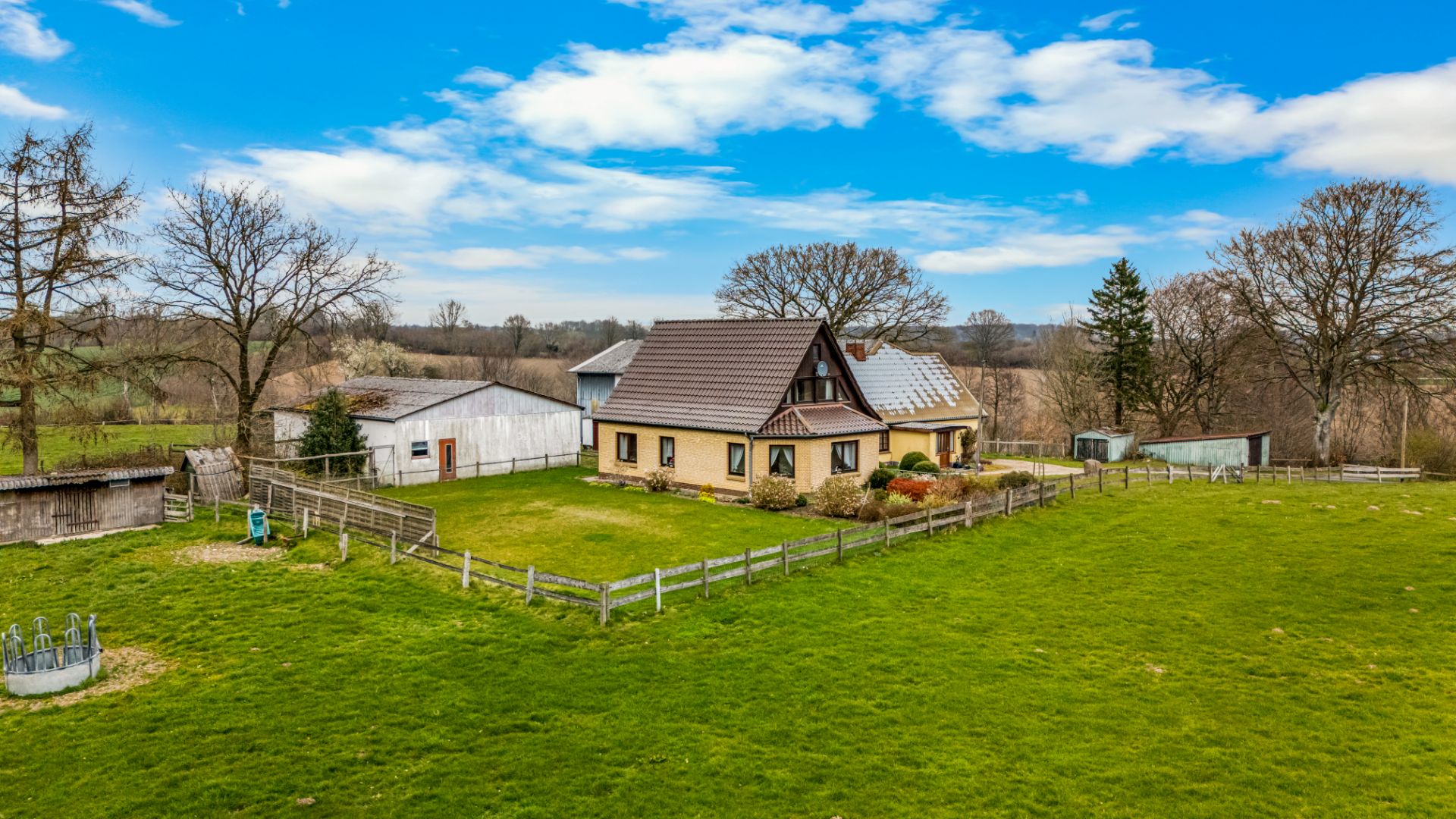 Einfamilienhaus mit Garten, umzäuntem Grundstück und Nebengebäuden auf großer Wiese.
