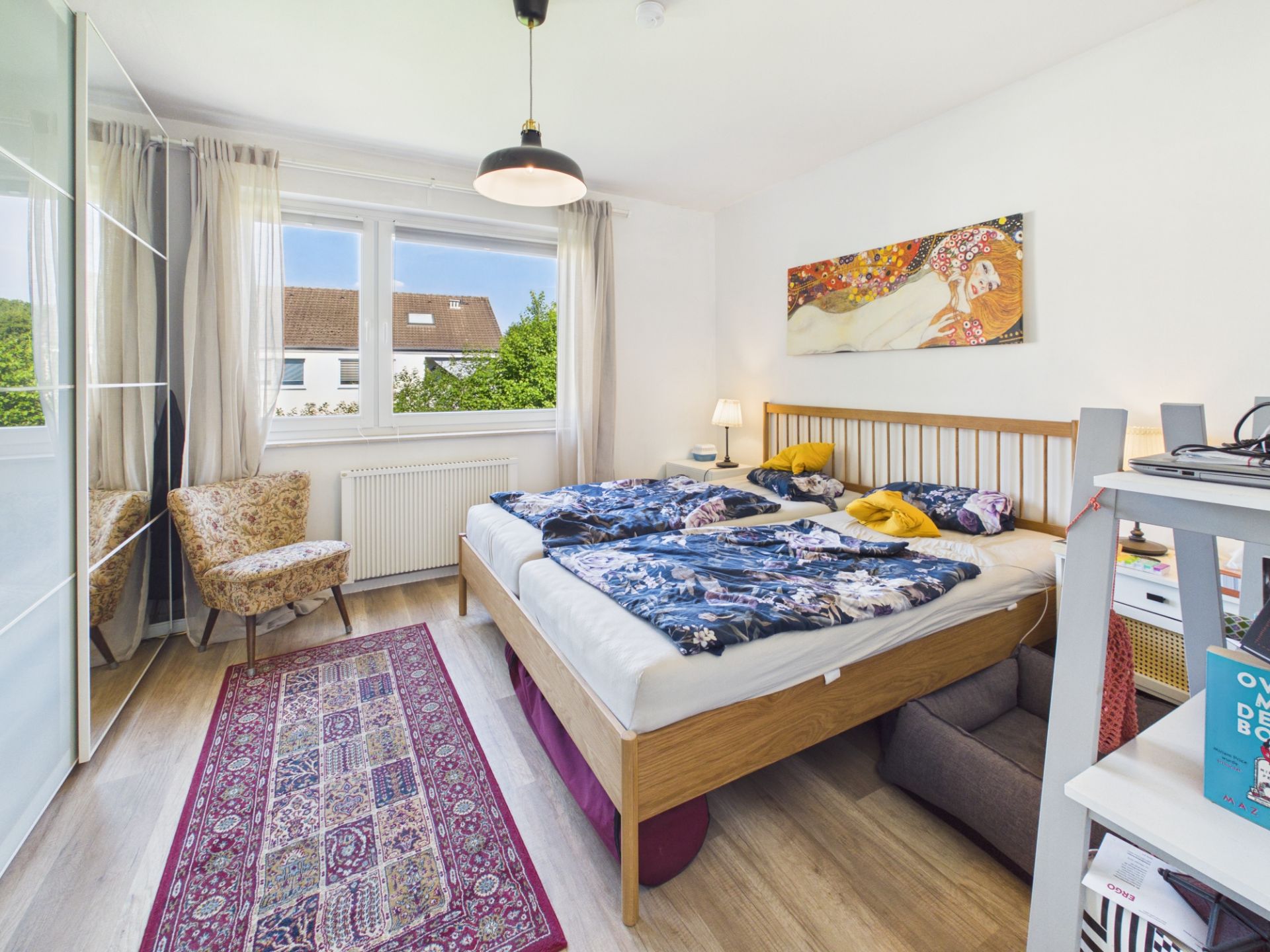 Helles Schlafzimmer mit Doppelbett, Teppich, Sessel und großem Fenster.