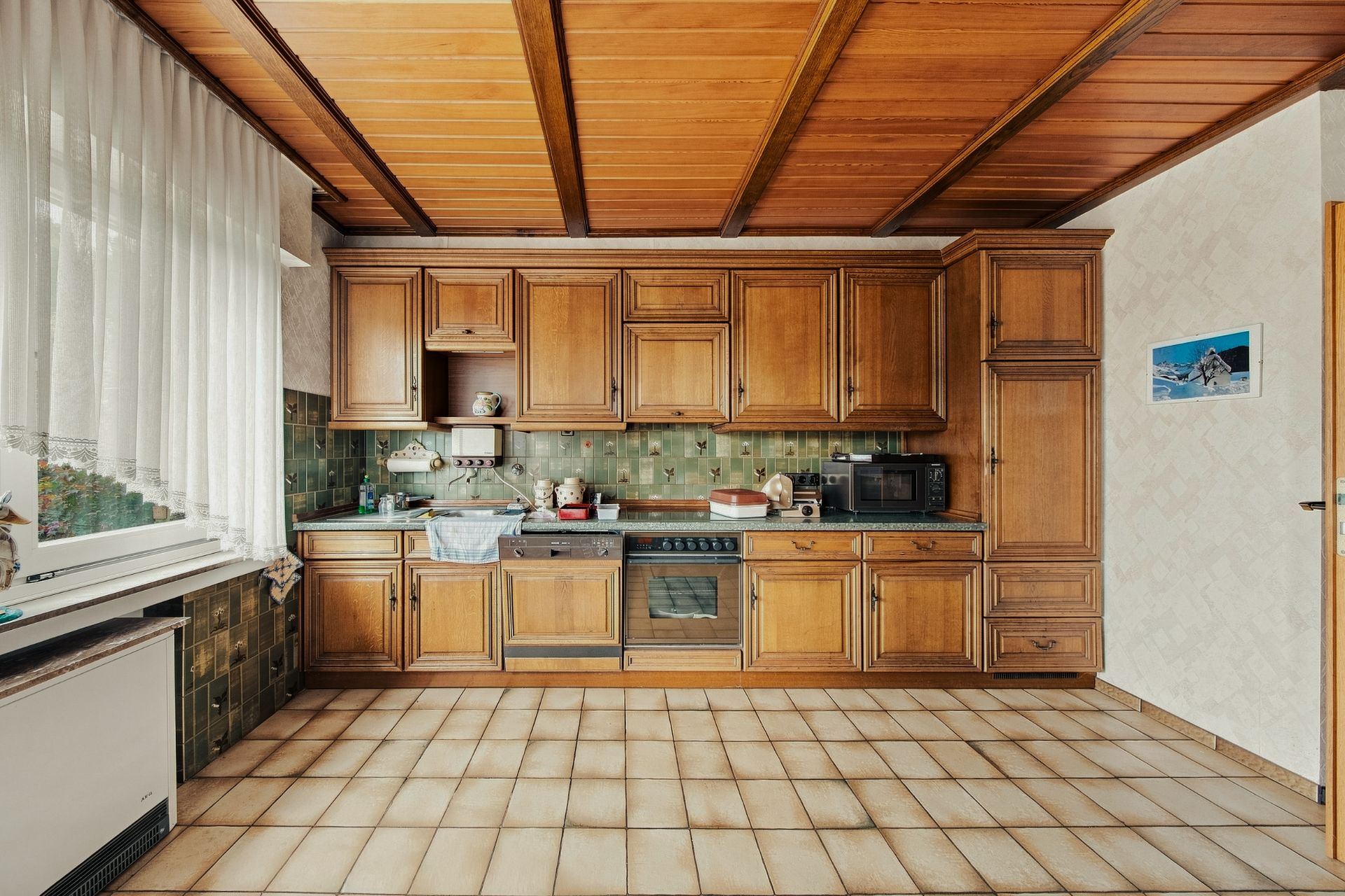 Küche mit Holzschränken, grünem Fliesenspiegel und großen Fenstern mit weißen Vorhängen.
