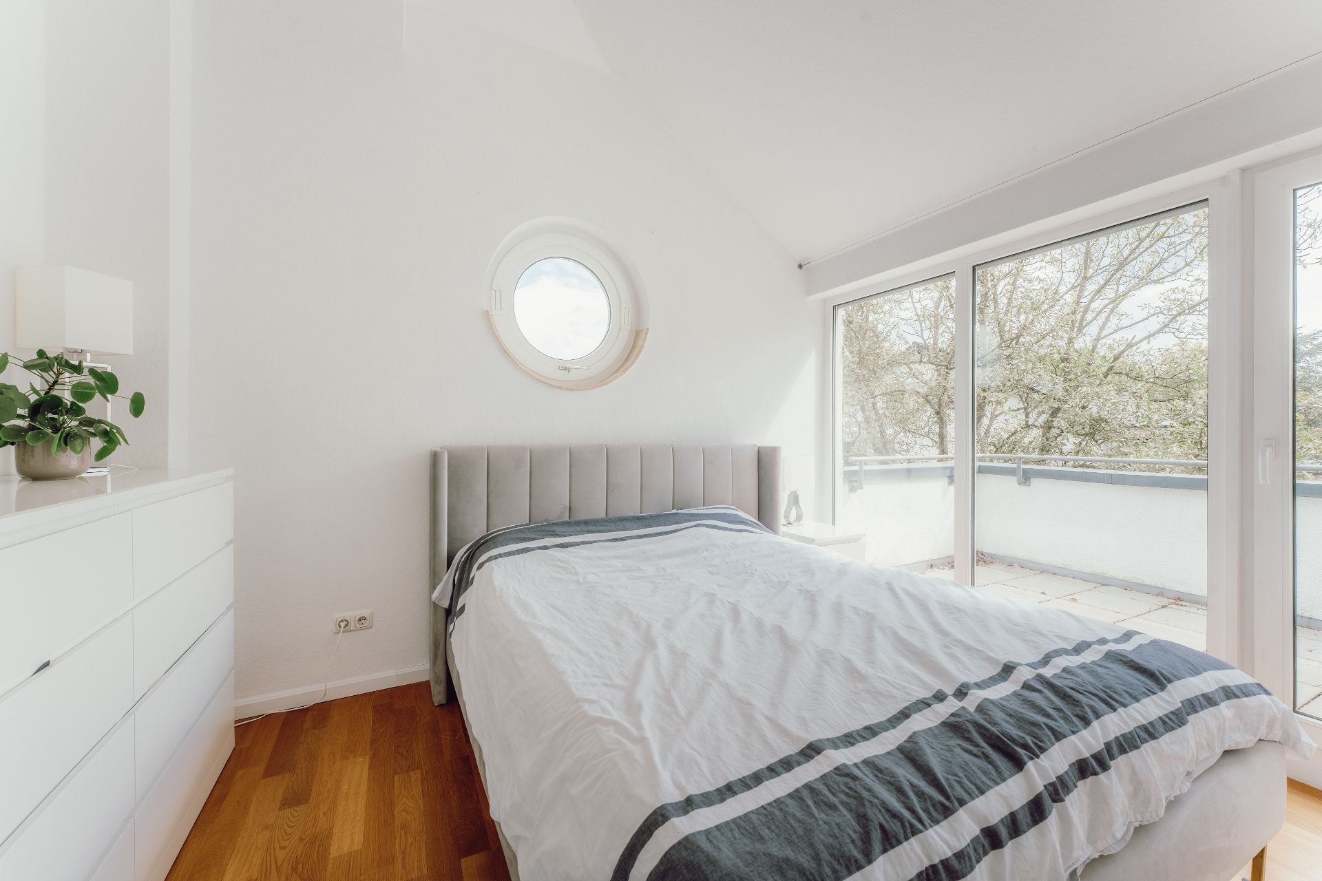 Helles Schlafzimmer mit Bett, Kommode, Rundfenster und Zugang zu einem Balkon.