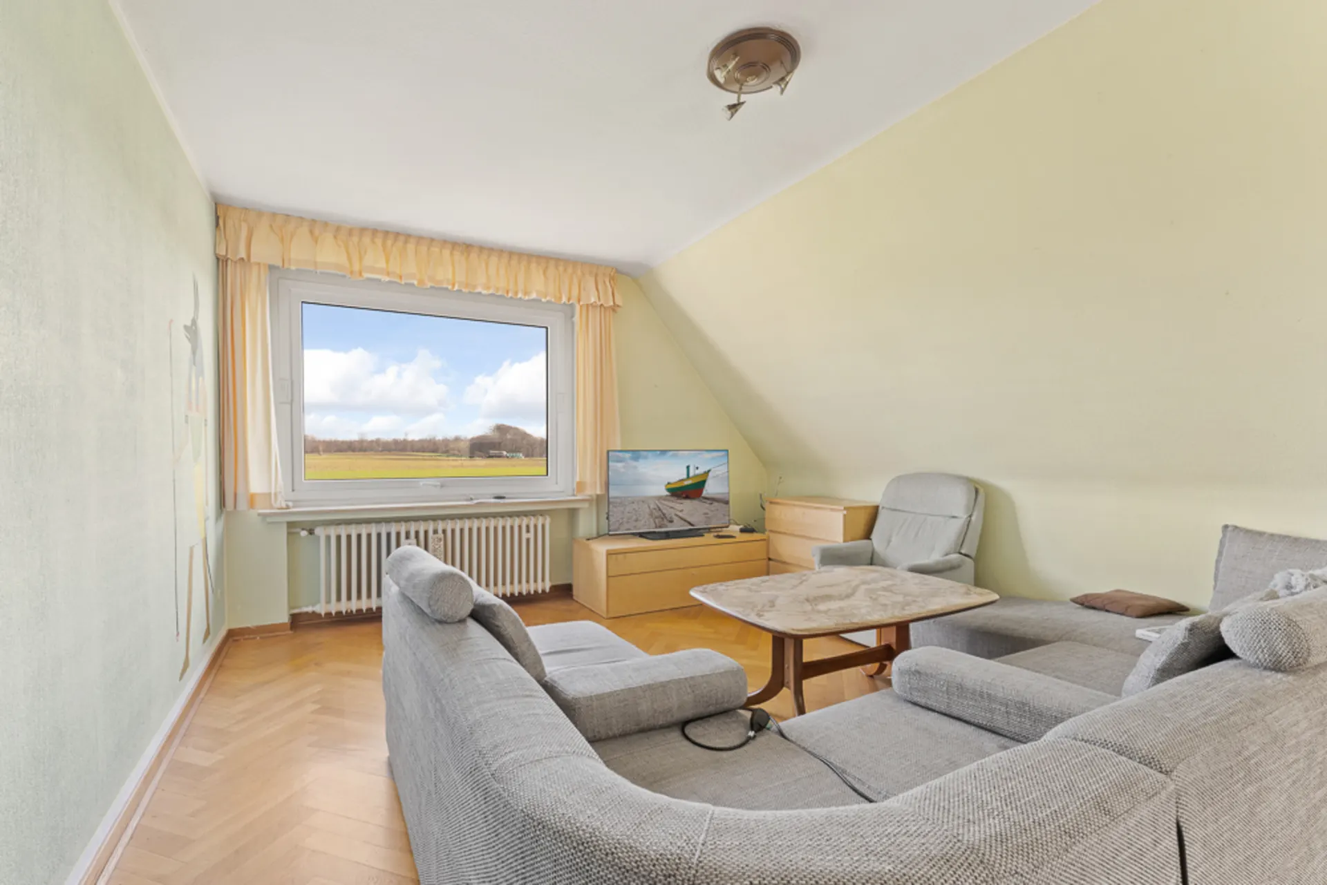 Wohnzimmer mit grauem Sofa, Sessel, Fernseher und großem Fenster mit Blick auf eine Wiese.